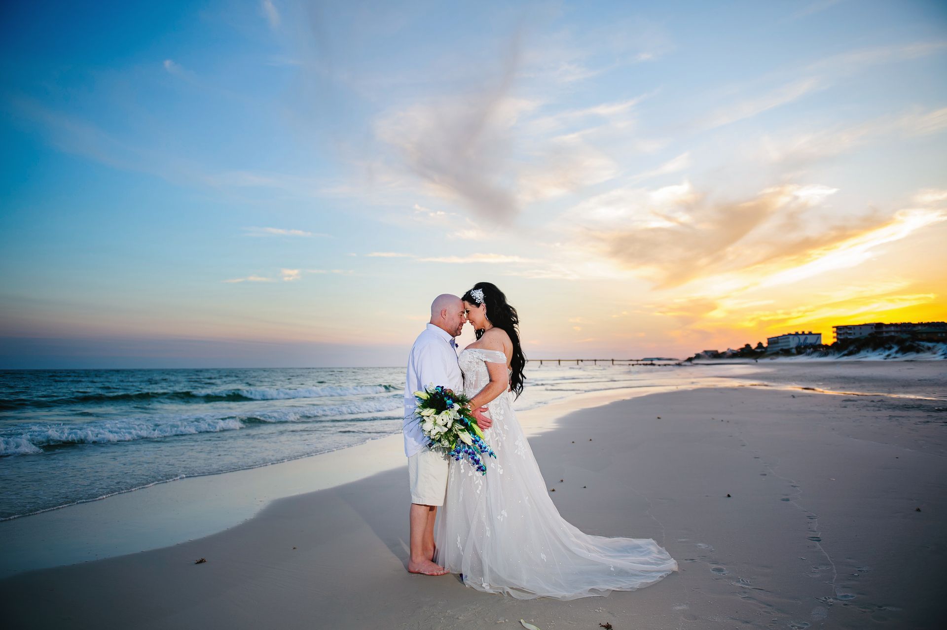 Wedding couple embracing on a beach at sunset, with ocean waves and a long white dress trailing on the sand