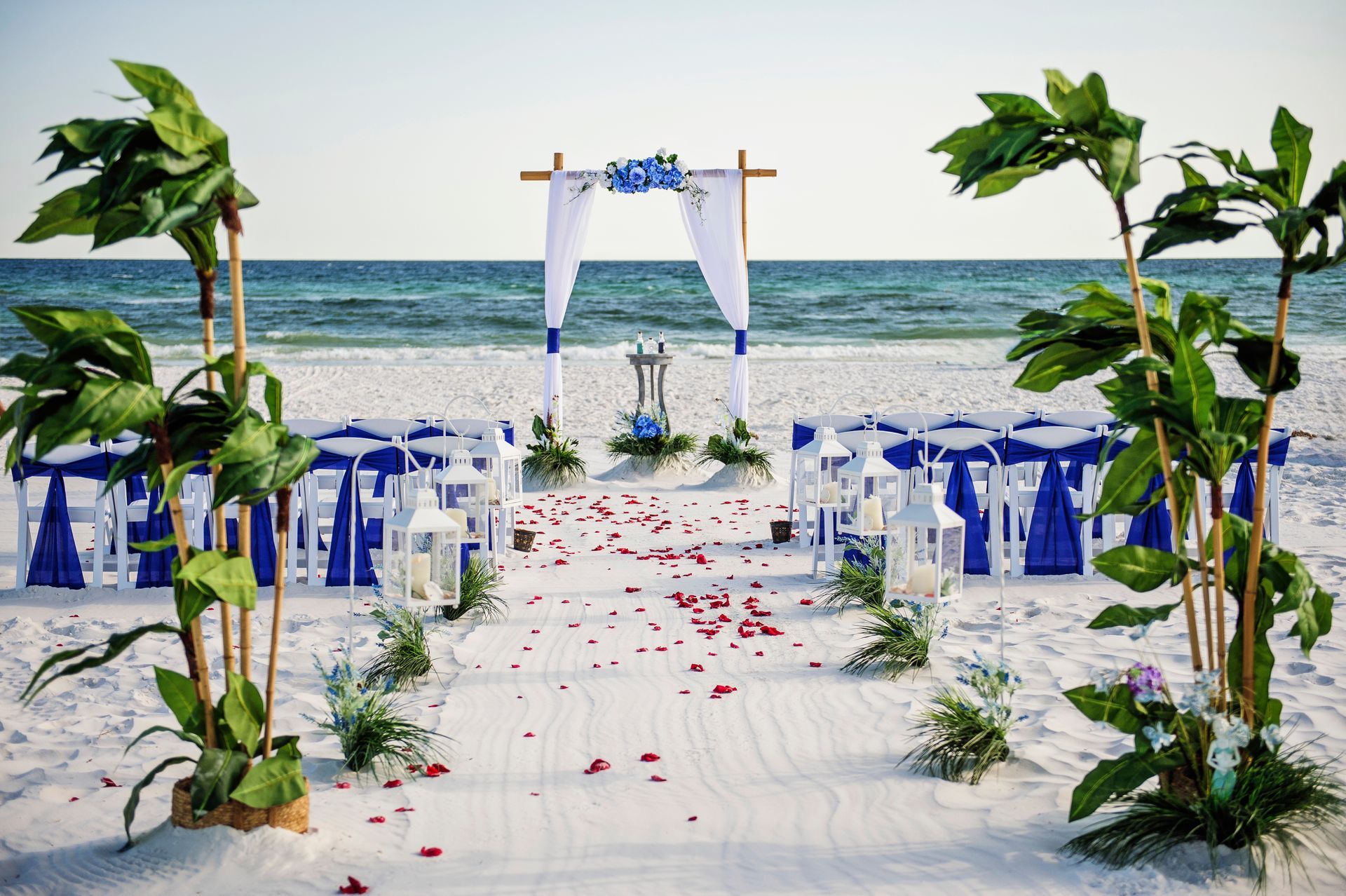 Beach wedding aisle with white chairs, blue ribbons, flower petals, and ocean backdrop
