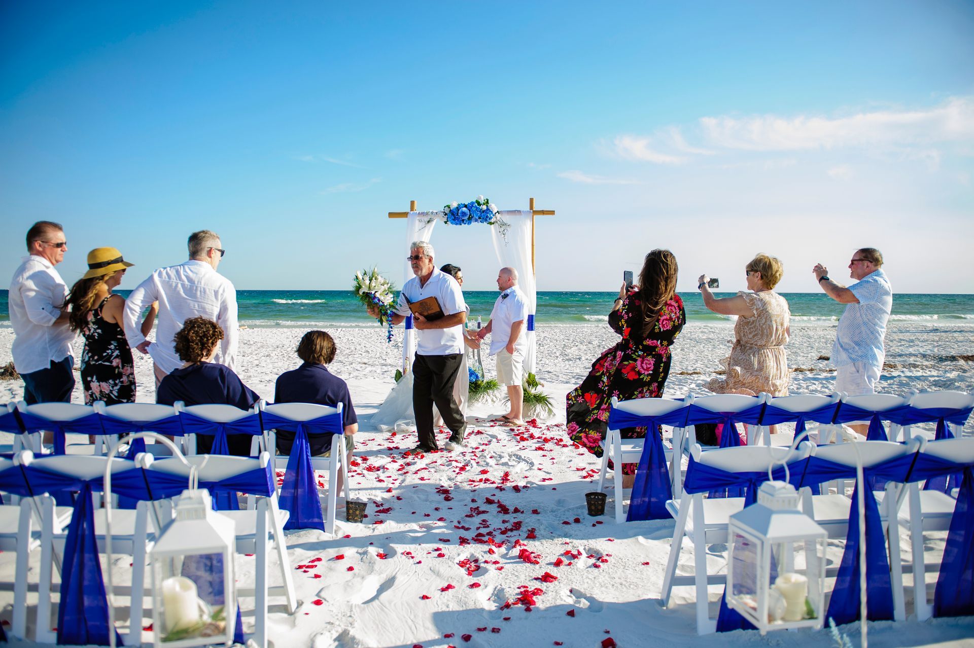 Beach wedding ceremony with guests seated on white and blue chairs, red petals on the aisle, and ocean backdrop