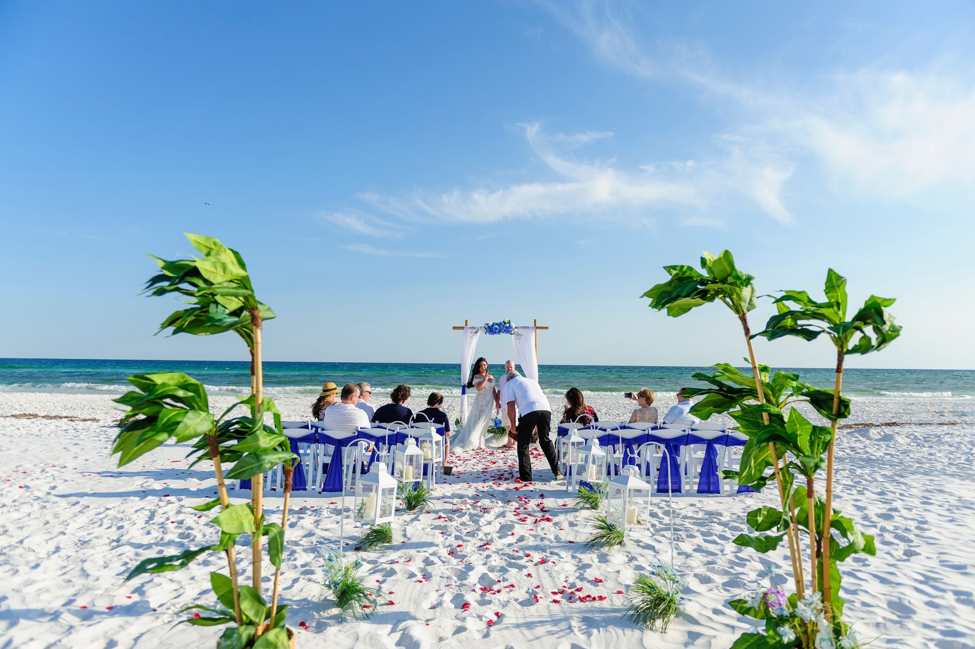 Beach wedding ceremony setup with white chairs and guests on a sunny shoreline under a blue sky