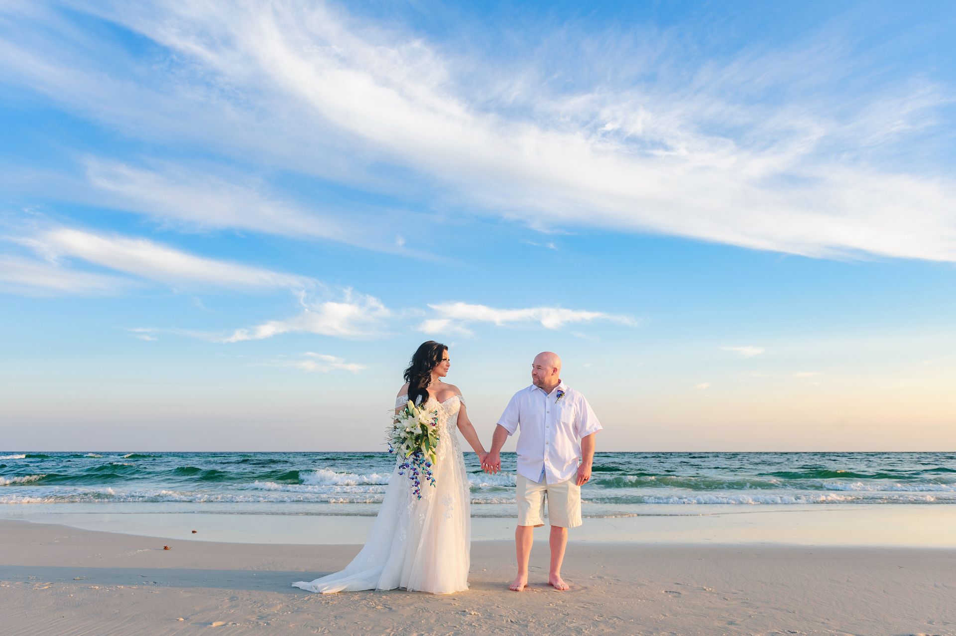 Couple holding hands on a beach at sunset, with waves and blue sky behind them