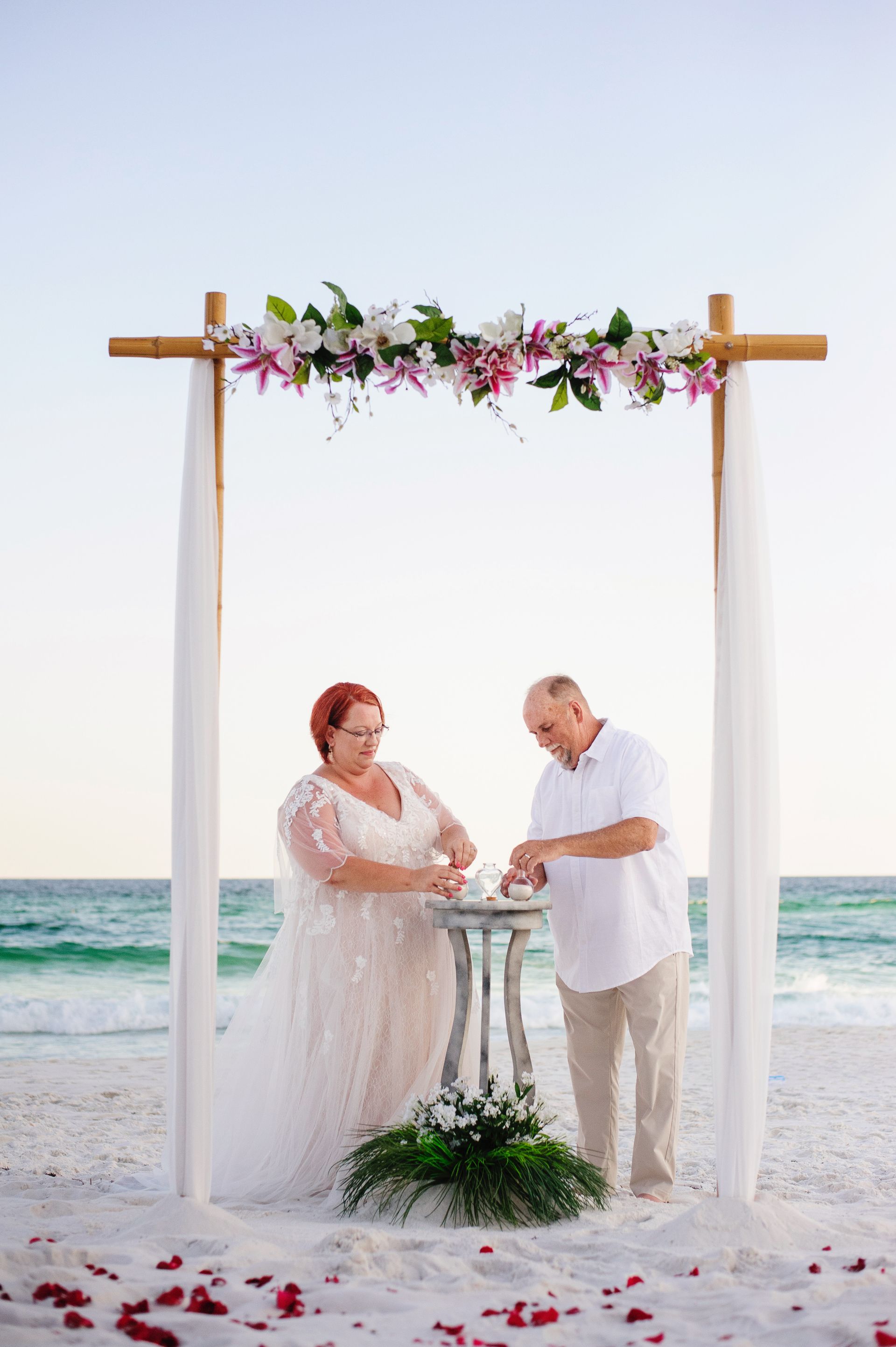 Couple exchanging vows under a floral arch on a beach at sunset