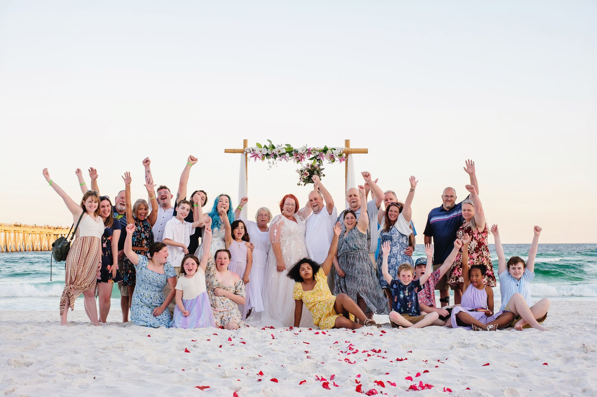Group of people celebrating on a beach at sunset, dancing and raising hands under a flower arch