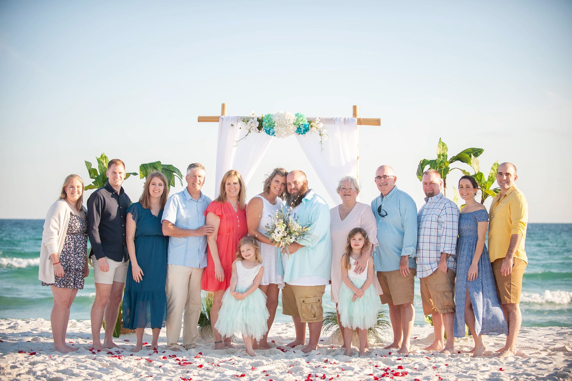 Group posing at a beach wedding arch, with bride and groom centered and guests smiling around them.