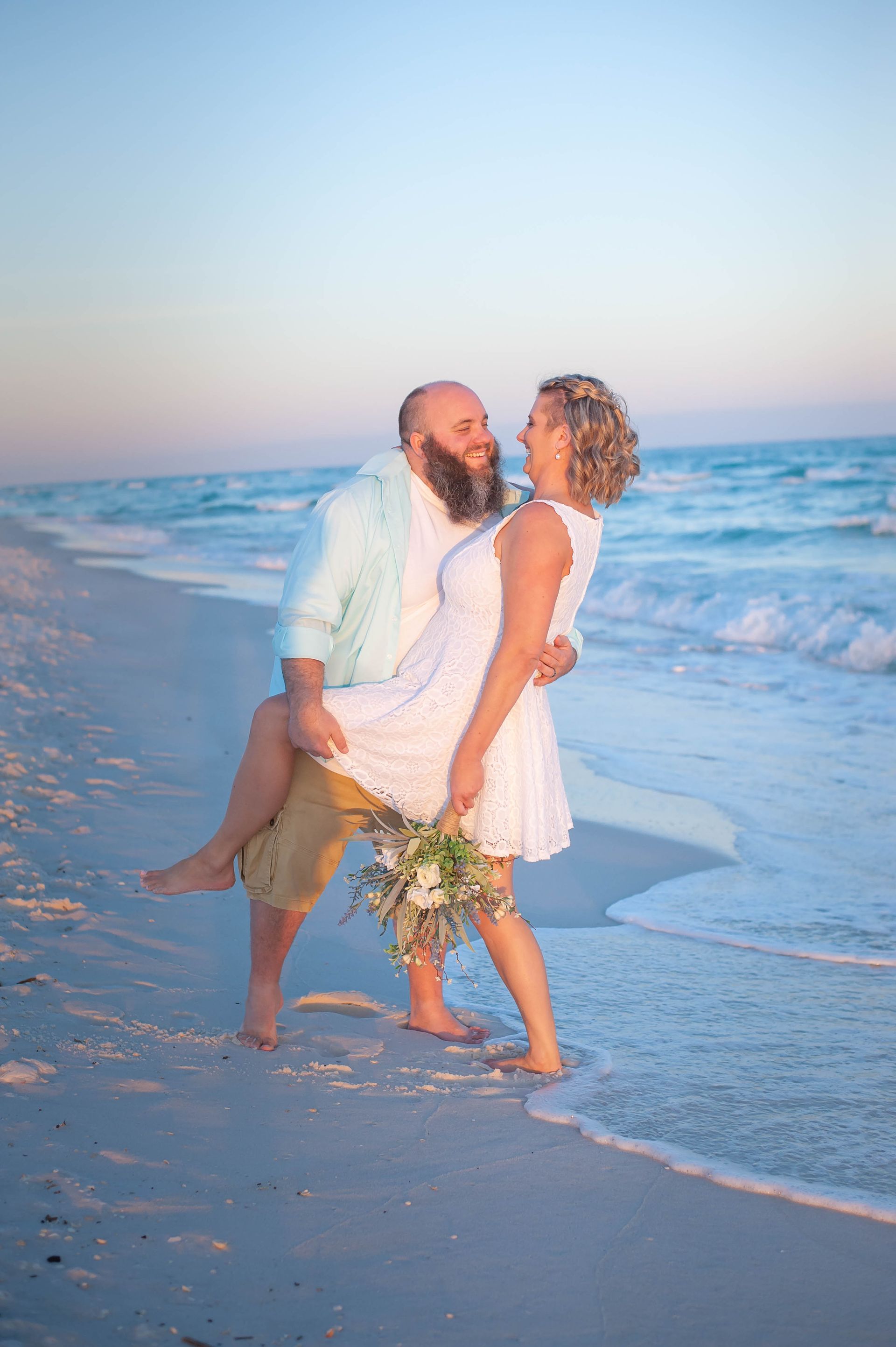 Couple walking barefoot on the beach at sunset, holding flowers and smiling by the ocean.