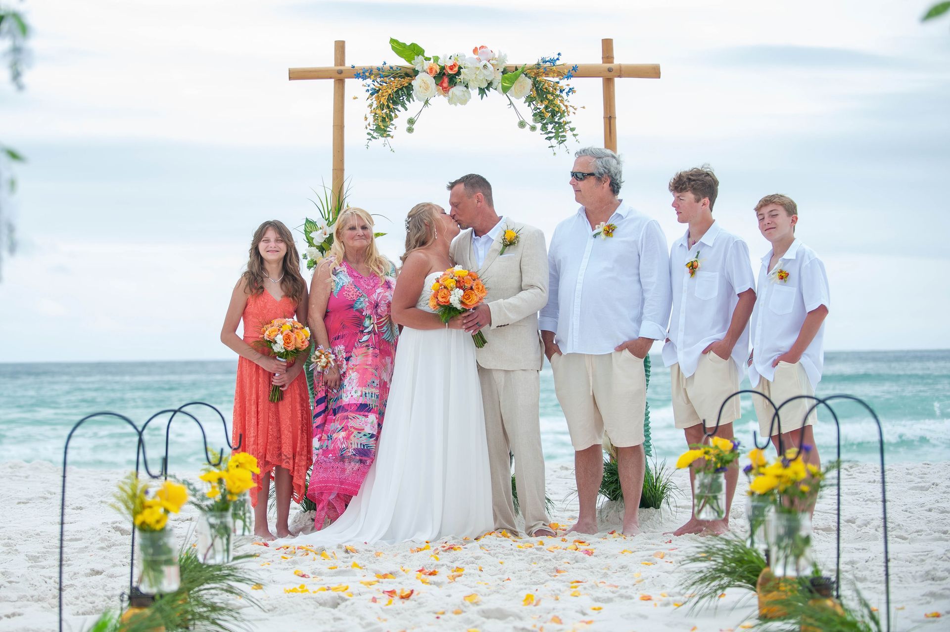 Wedding party posing on a dock by the sea under a floral arch, with the bride and groom at center.