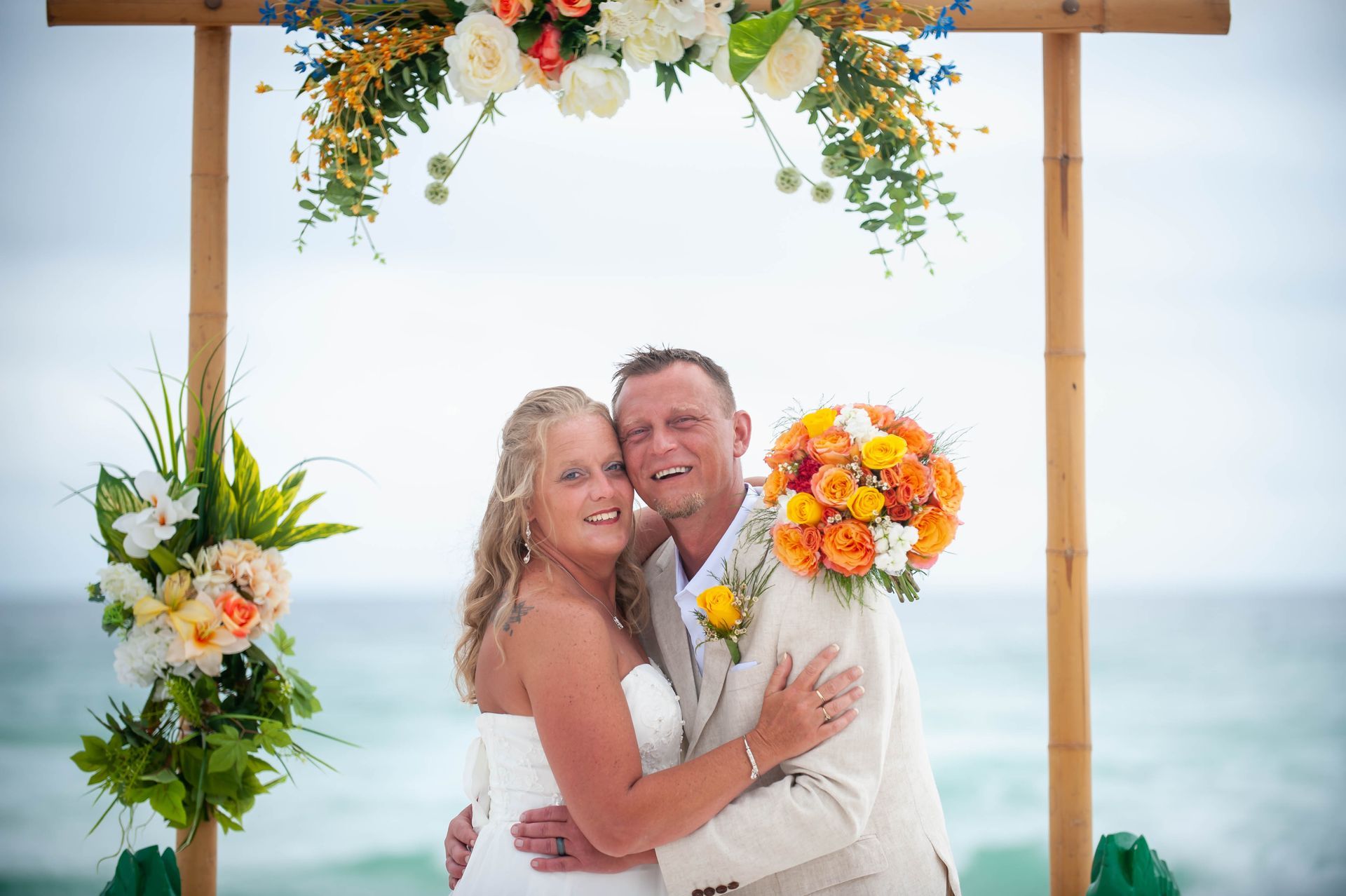 Couple embracing under a flower-decorated arch on a beach, smiling and holding a bouquet