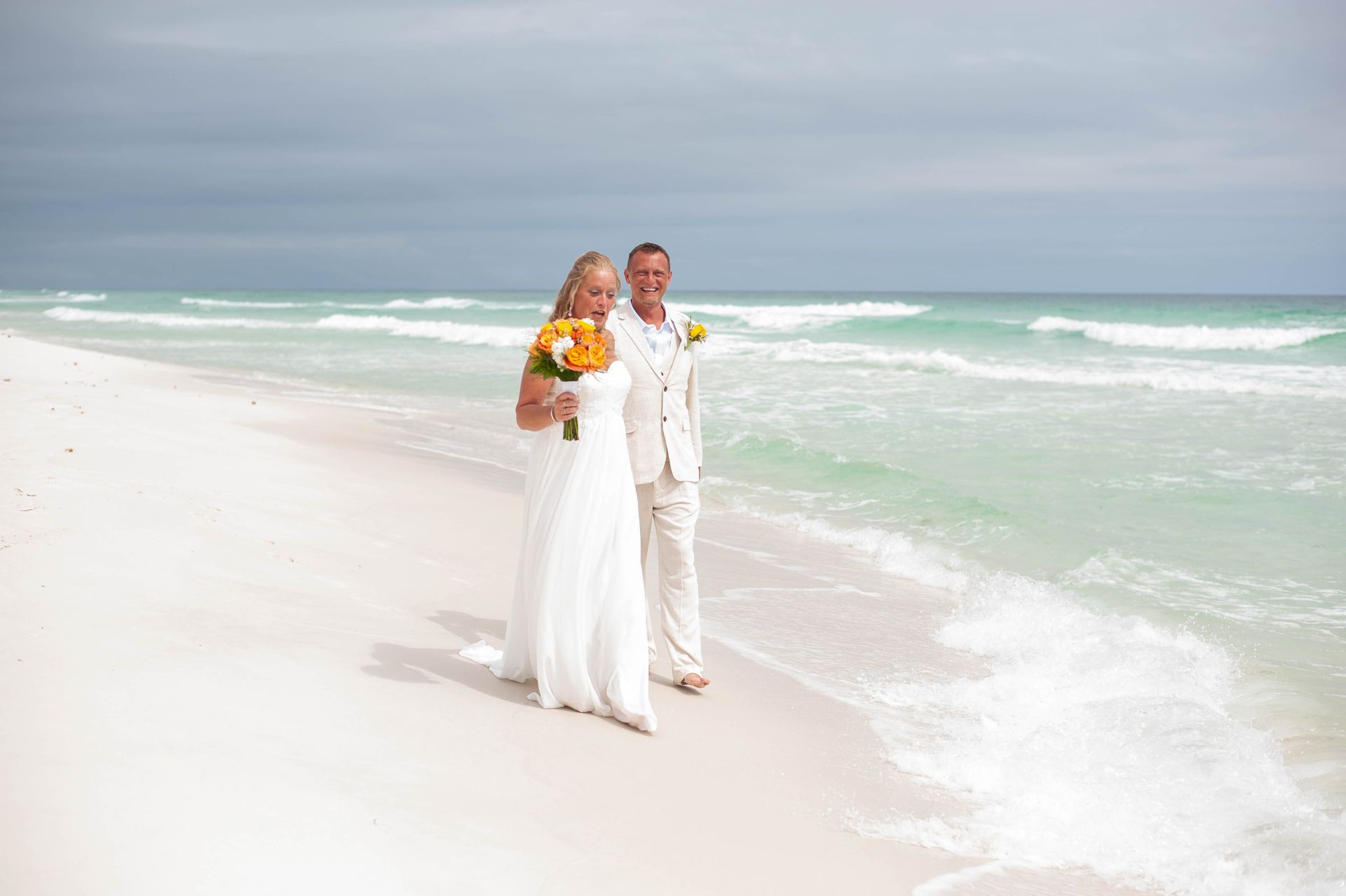 Couple in white standing on a bright beach beside turquoise waves, holding a bouquet