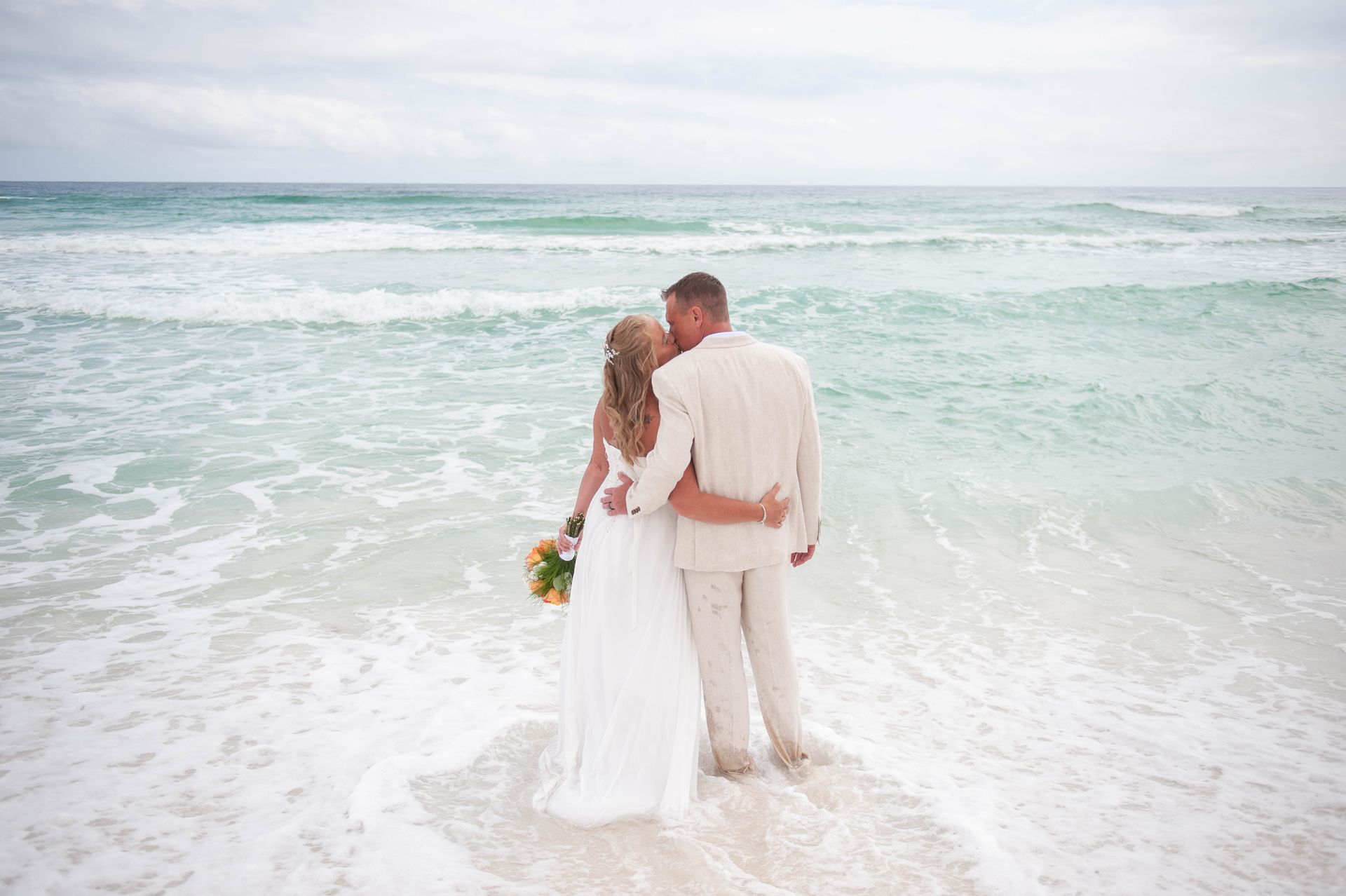 Couple in white clothes standing at the edge of the ocean, facing the waves on a beach.
