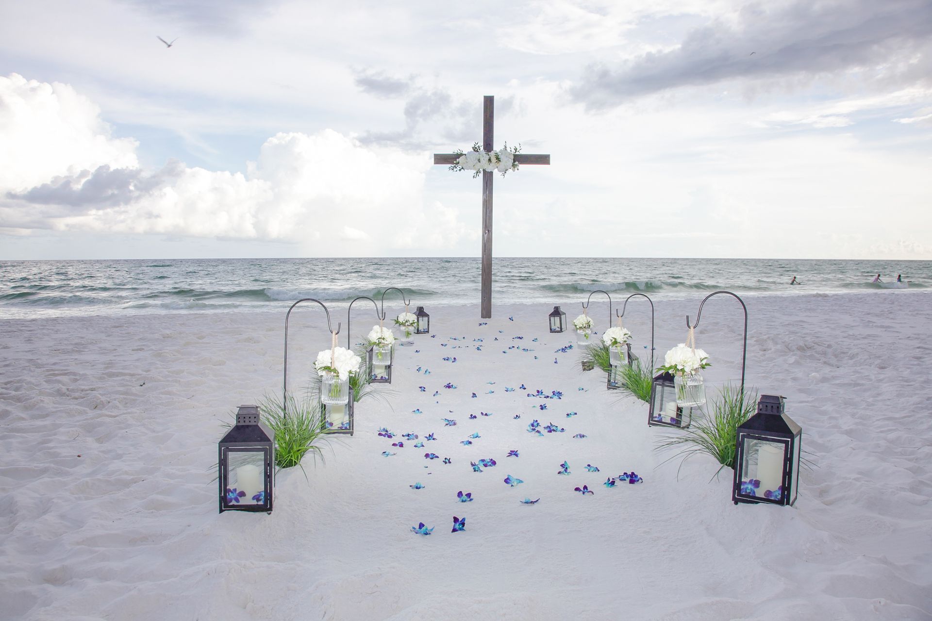 Beach wedding aisle with lanterns and blue petals leading to a cross by the ocean