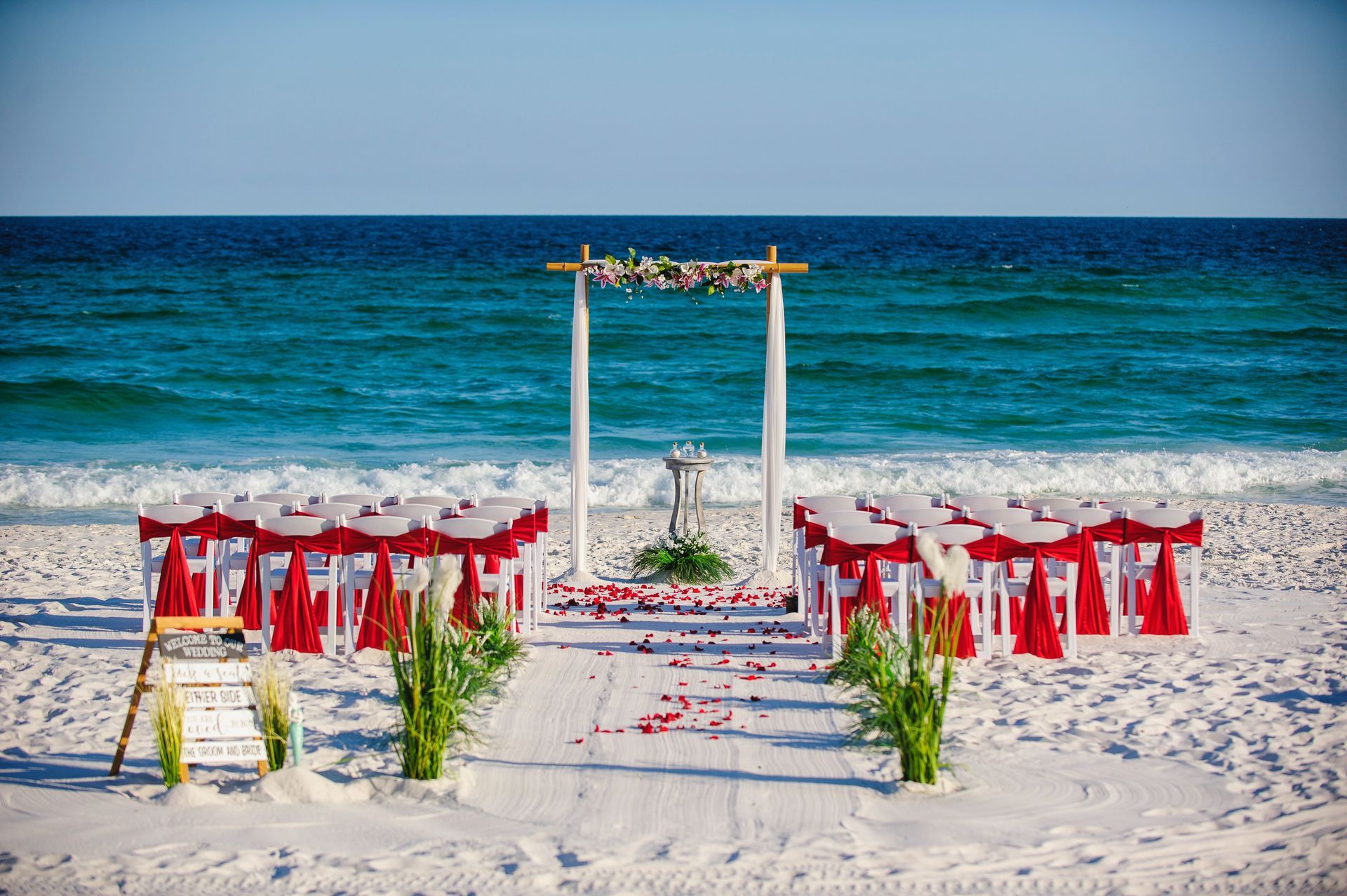Beach wedding setup with red-and-white chairs, white arch, and floral aisle by the ocean