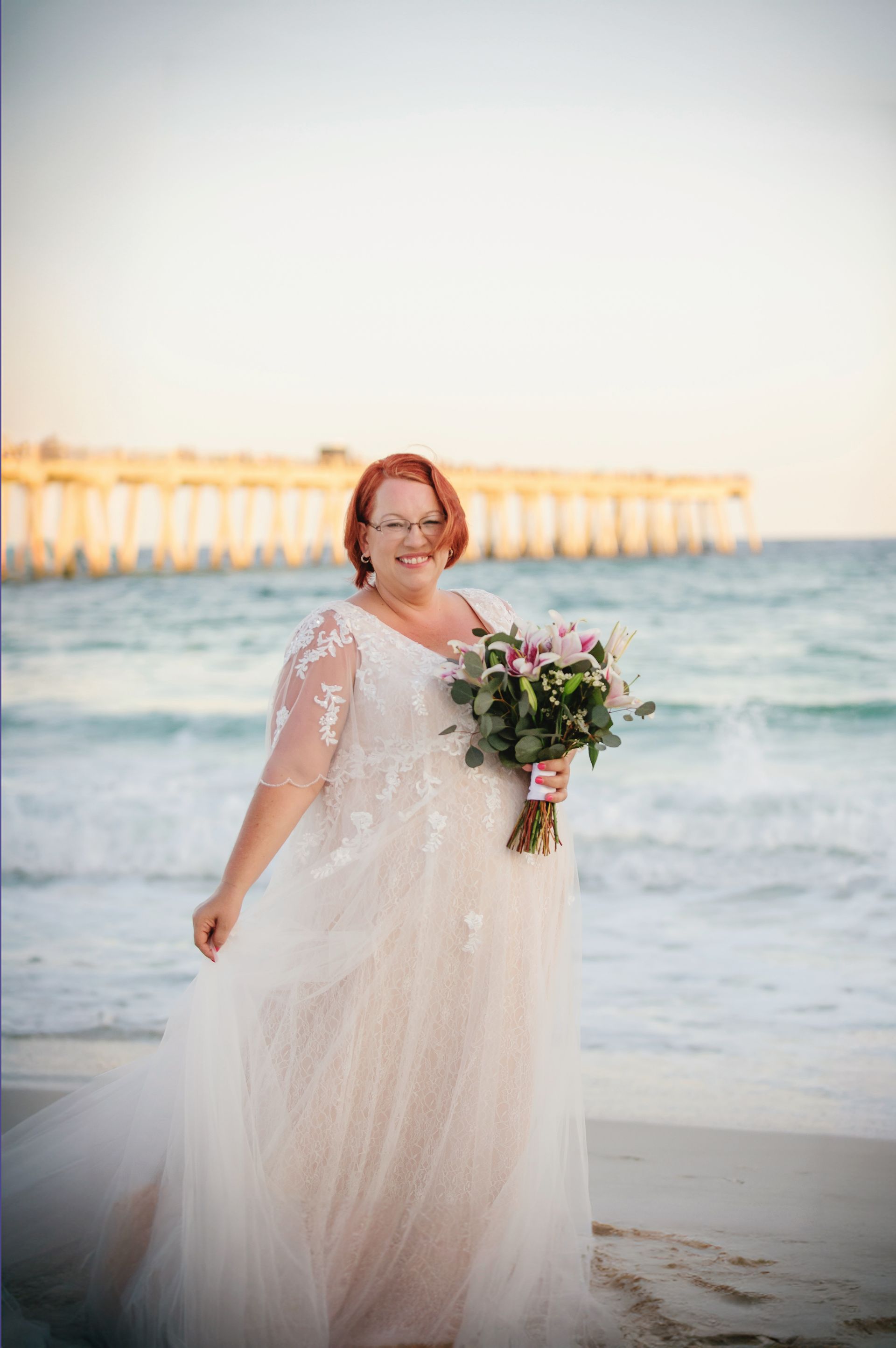 Bride in a white lace dress holding a bouquet on a beach at sunset