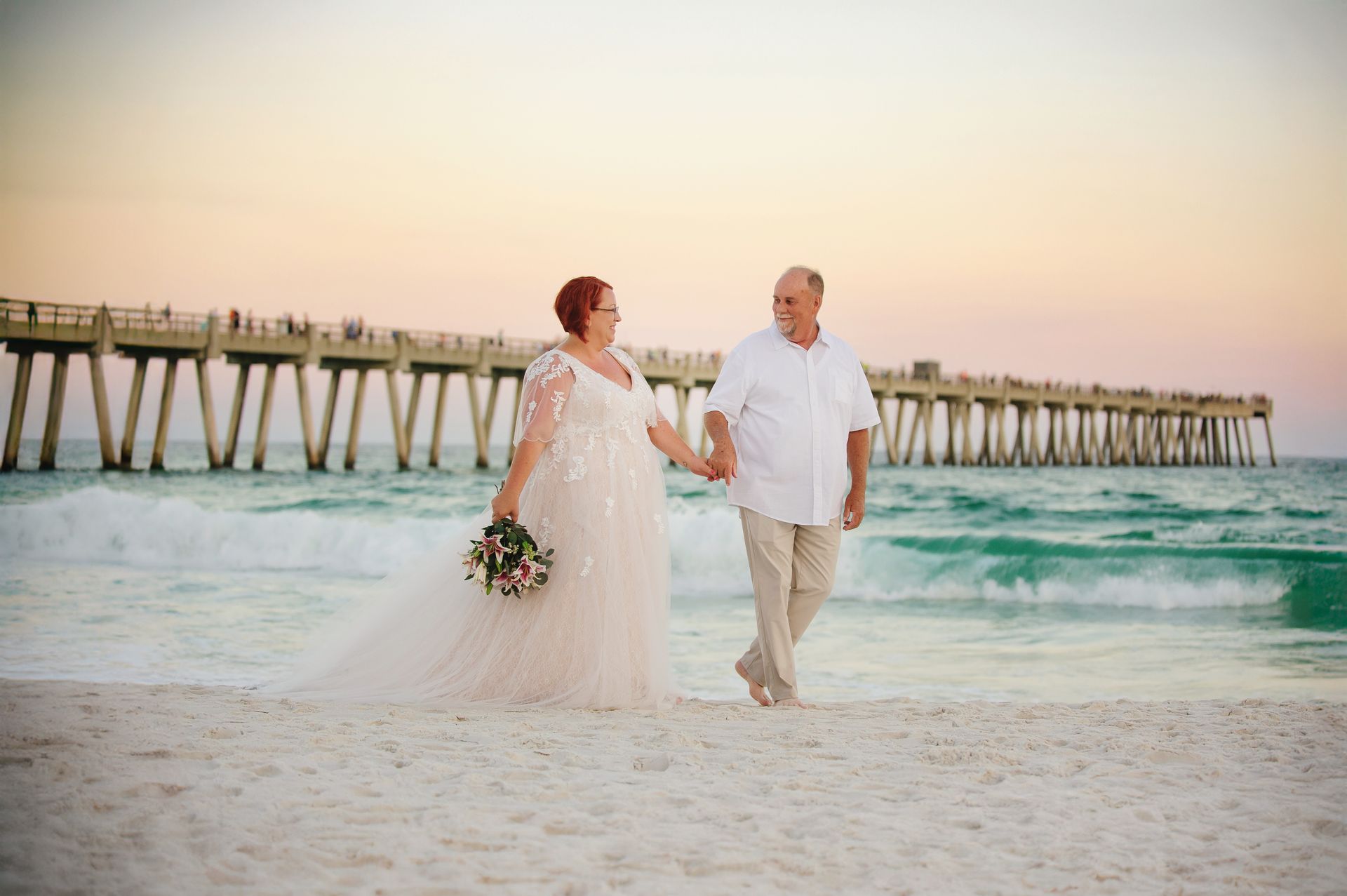 Couple walking hand in hand on a beach near a pier at sunset