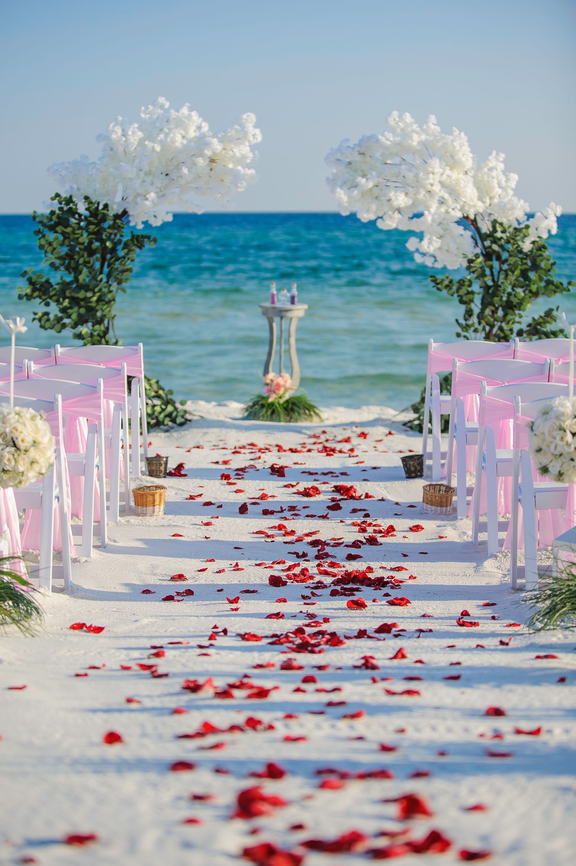 Rose-petal aisle to a beach altar framed by white flowers, with ocean waves beyond.