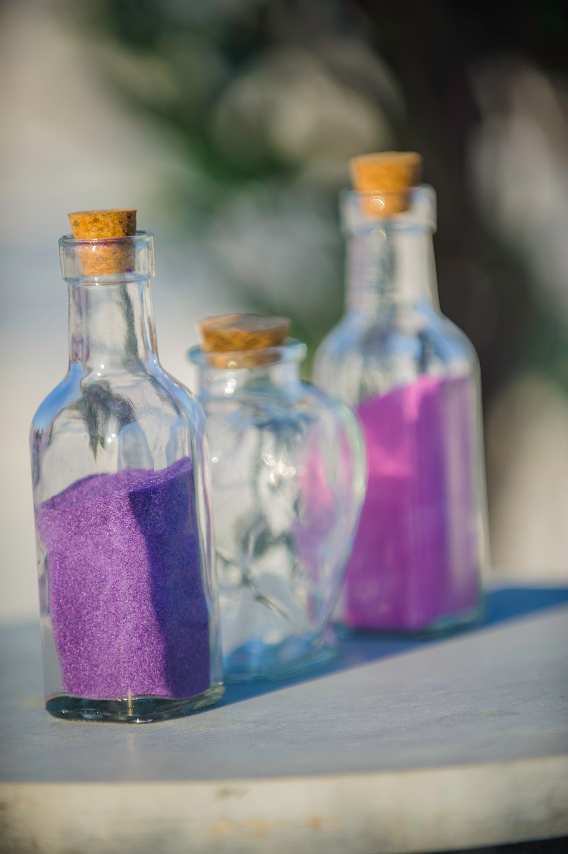 Three glass bottles with corks, filled with purple and clear liquid, on a table outdoors