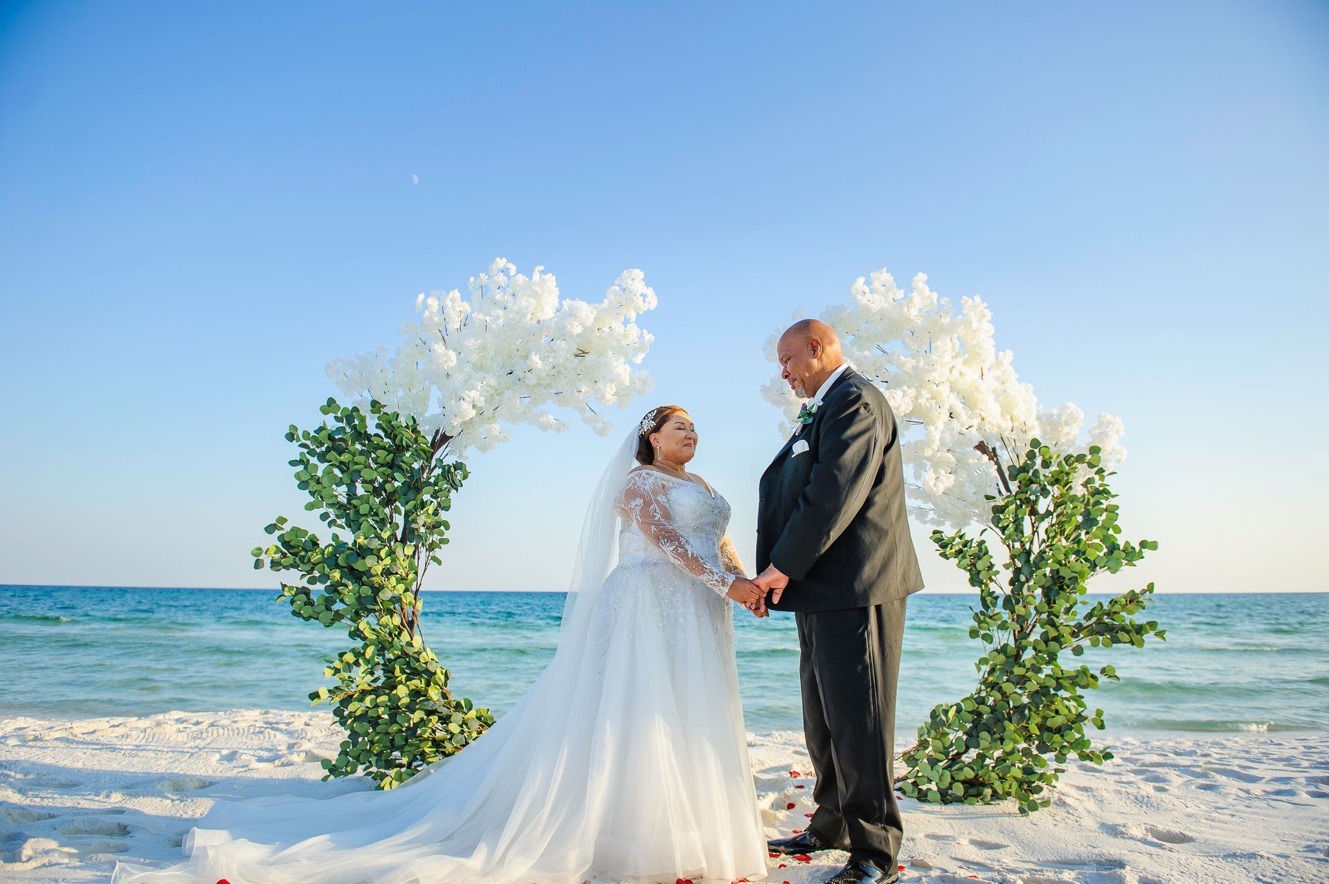 Bride and groom standing on a beach under white floral arch, facing each other in the sun