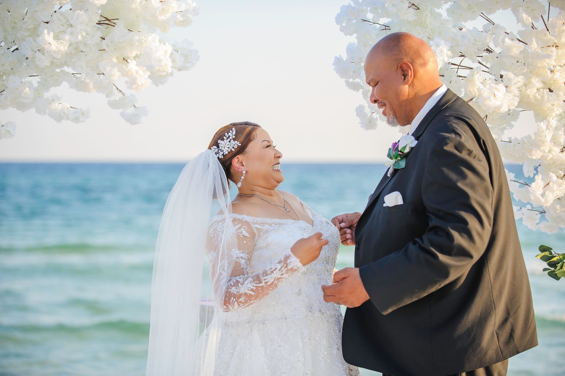 Bride and groom holding hands on a beach under flowering trees, smiling at each other