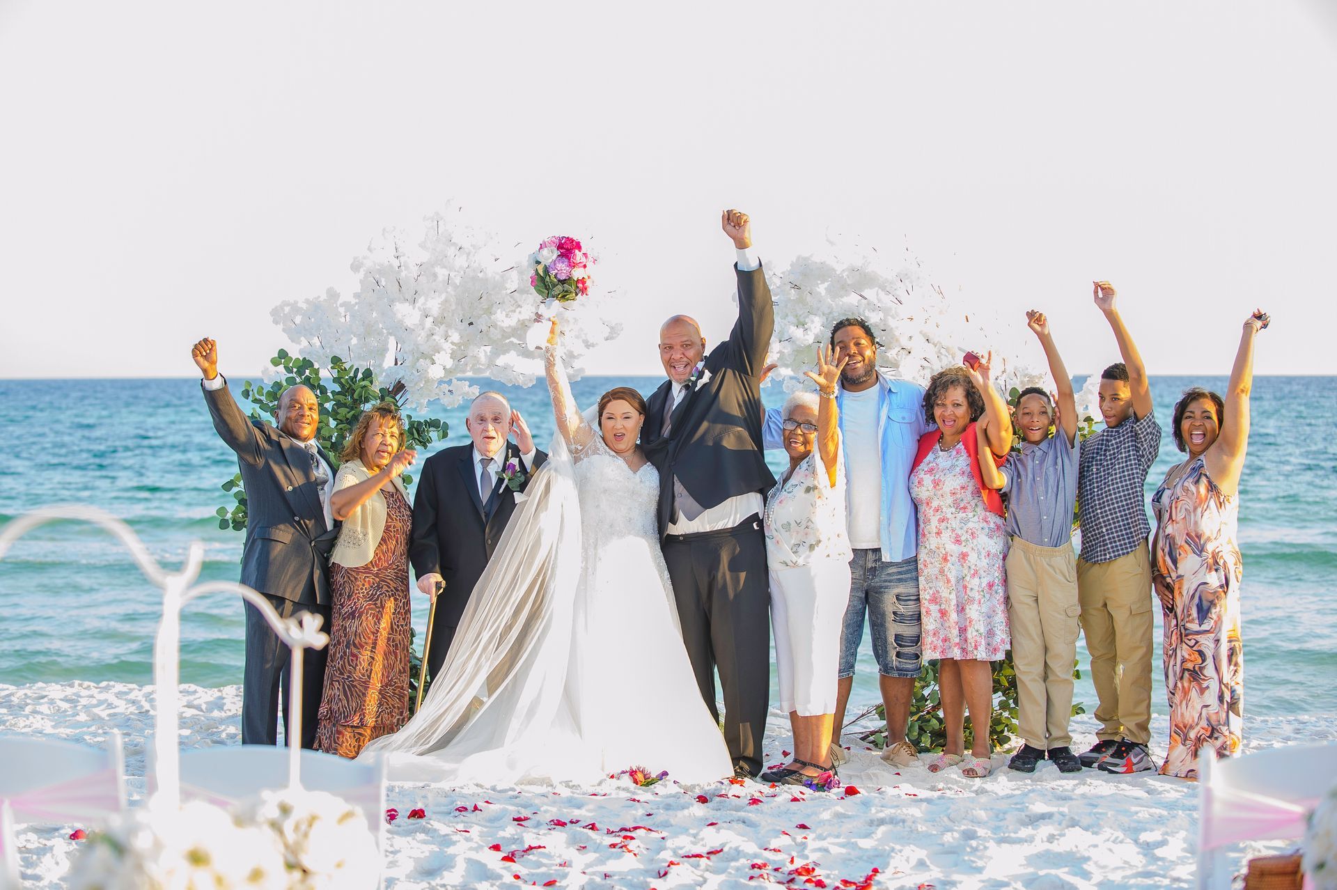 Wedding party celebrating on a boat deck, newlyweds centered with guests raising their arms by the ocean.