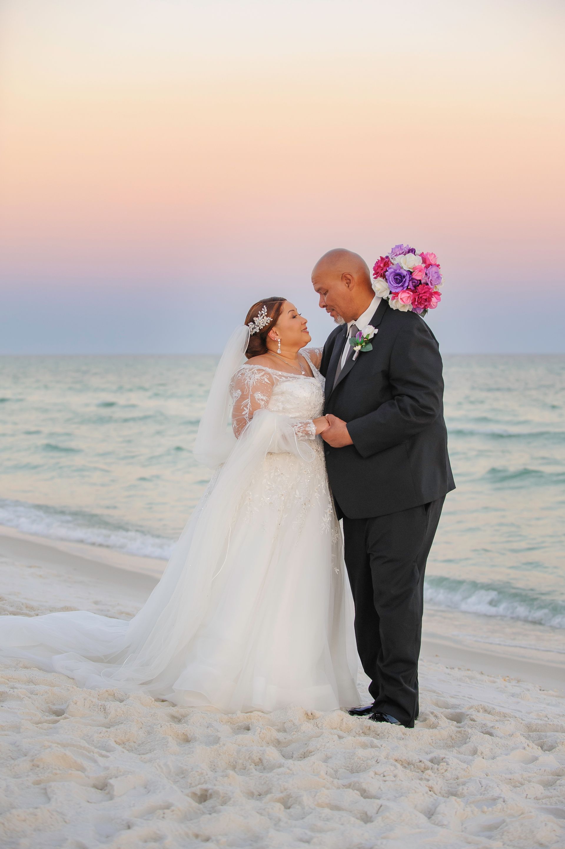 Newlywed couple embracing on a sandy beach at sunset, with pastel sky and ocean waves behind them