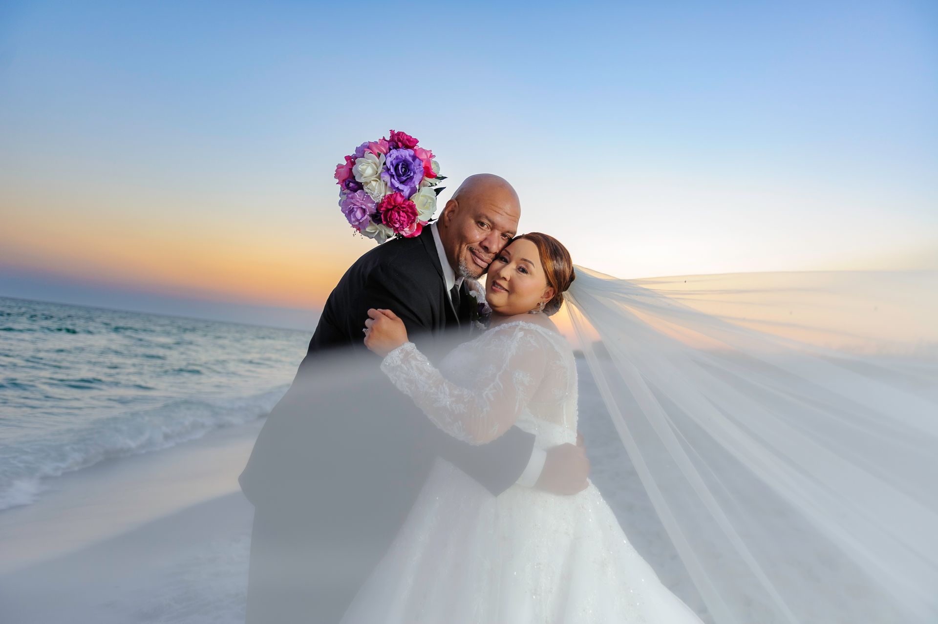 Couple embracing on a beach at sunset, bride in white gown with veil and groom in black suit