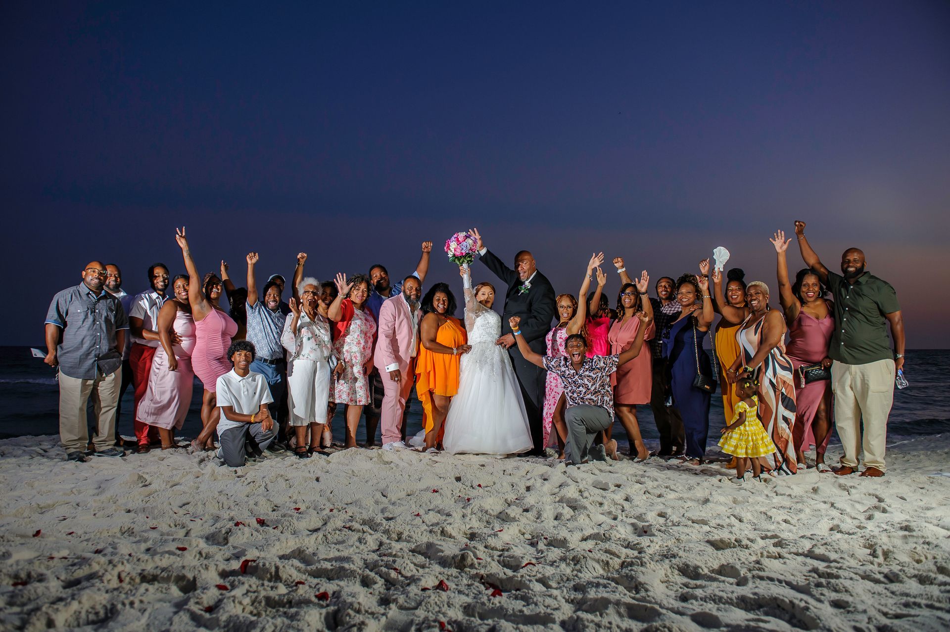 Large group posing on a beach at dusk, many waving under a dark blue sky, with a bride in white at center