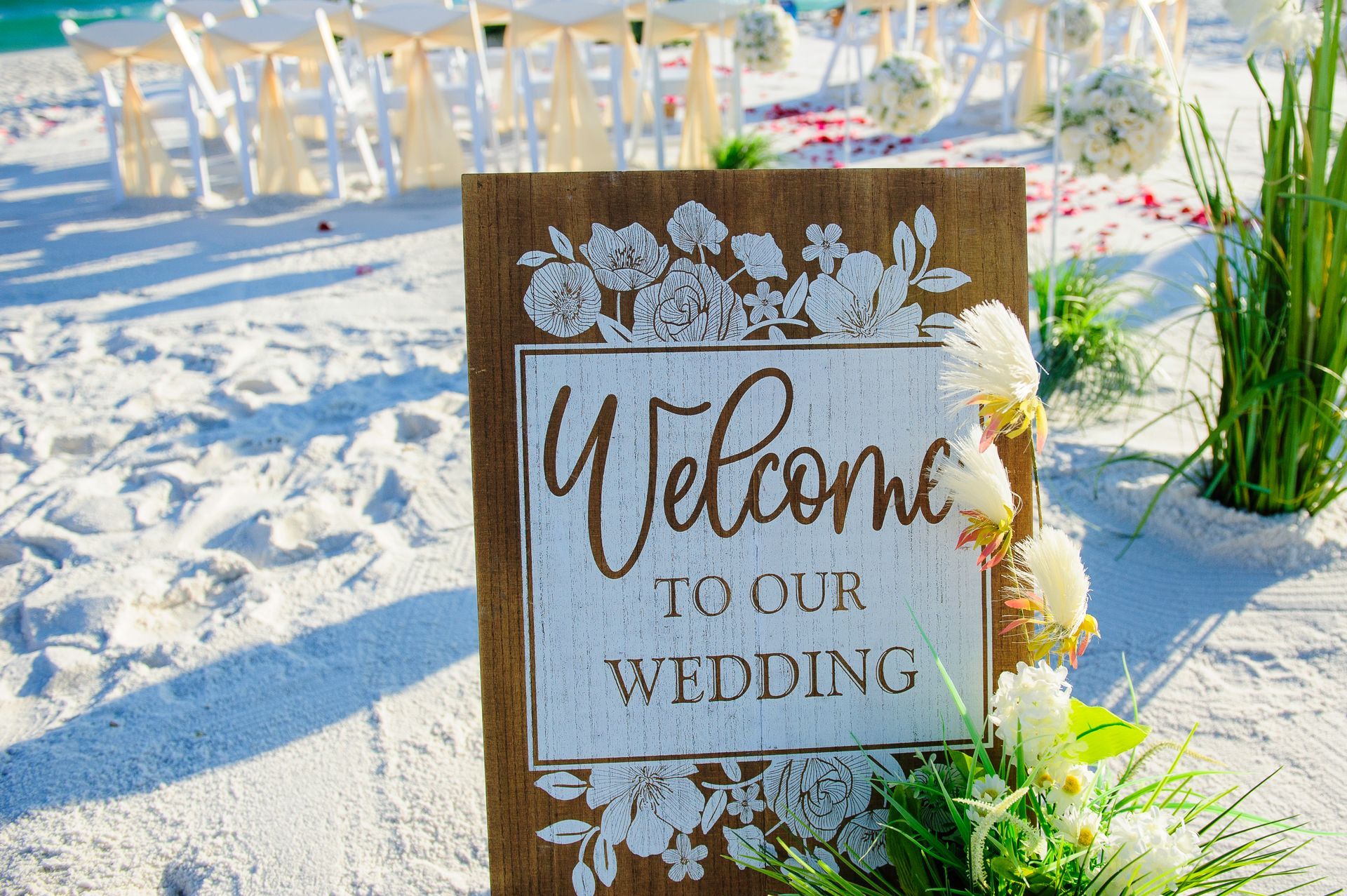 Wooden “Welcome to our wedding” sign on a sandy beach with white floral decor and outdoor tables nearby