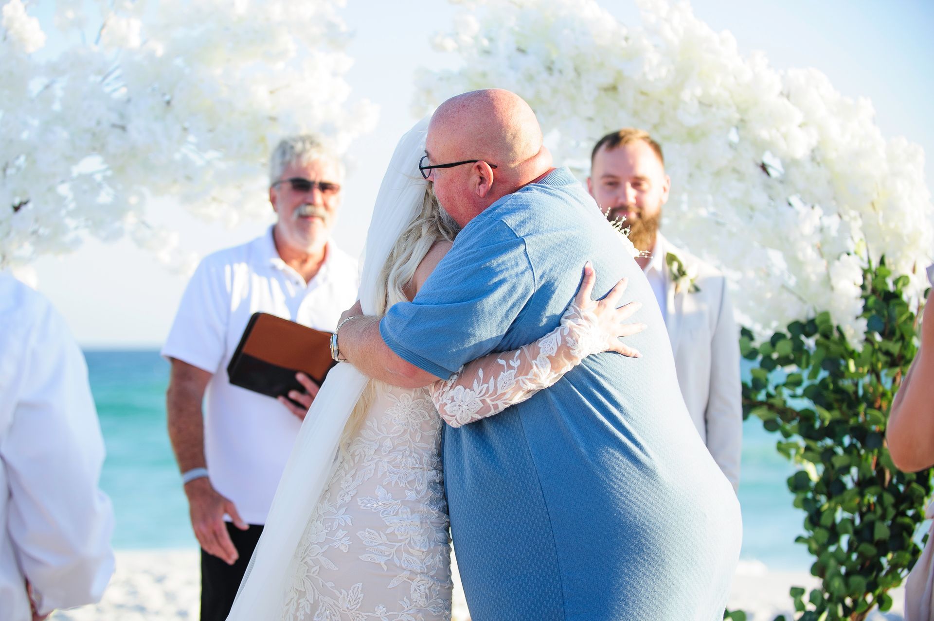 Bride and groom embracing at an outdoor wedding ceremony, with an officiant and white floral arch in the background