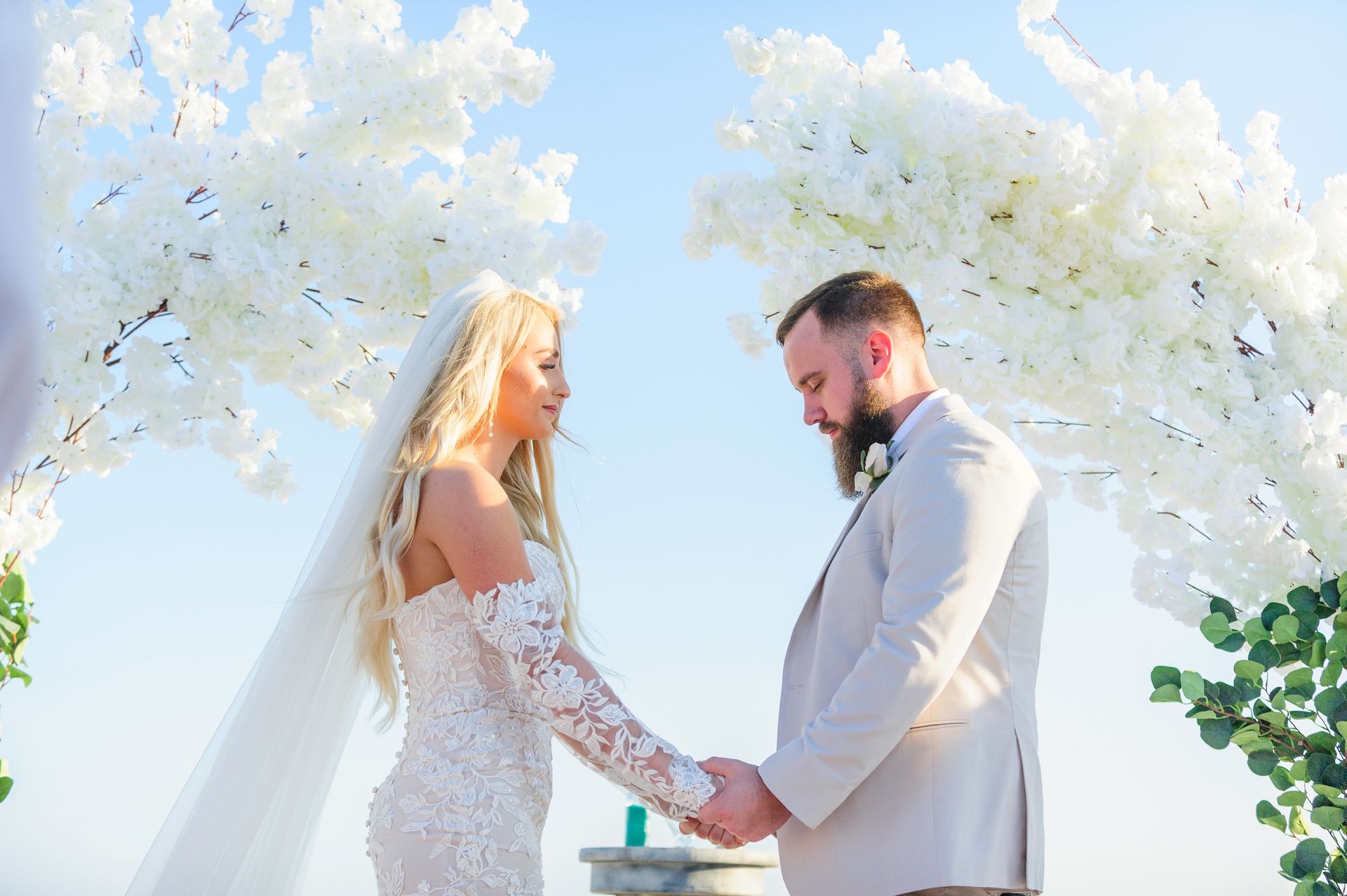 Bride and groom holding hands beneath white floral arches outdoors at a wedding ceremony