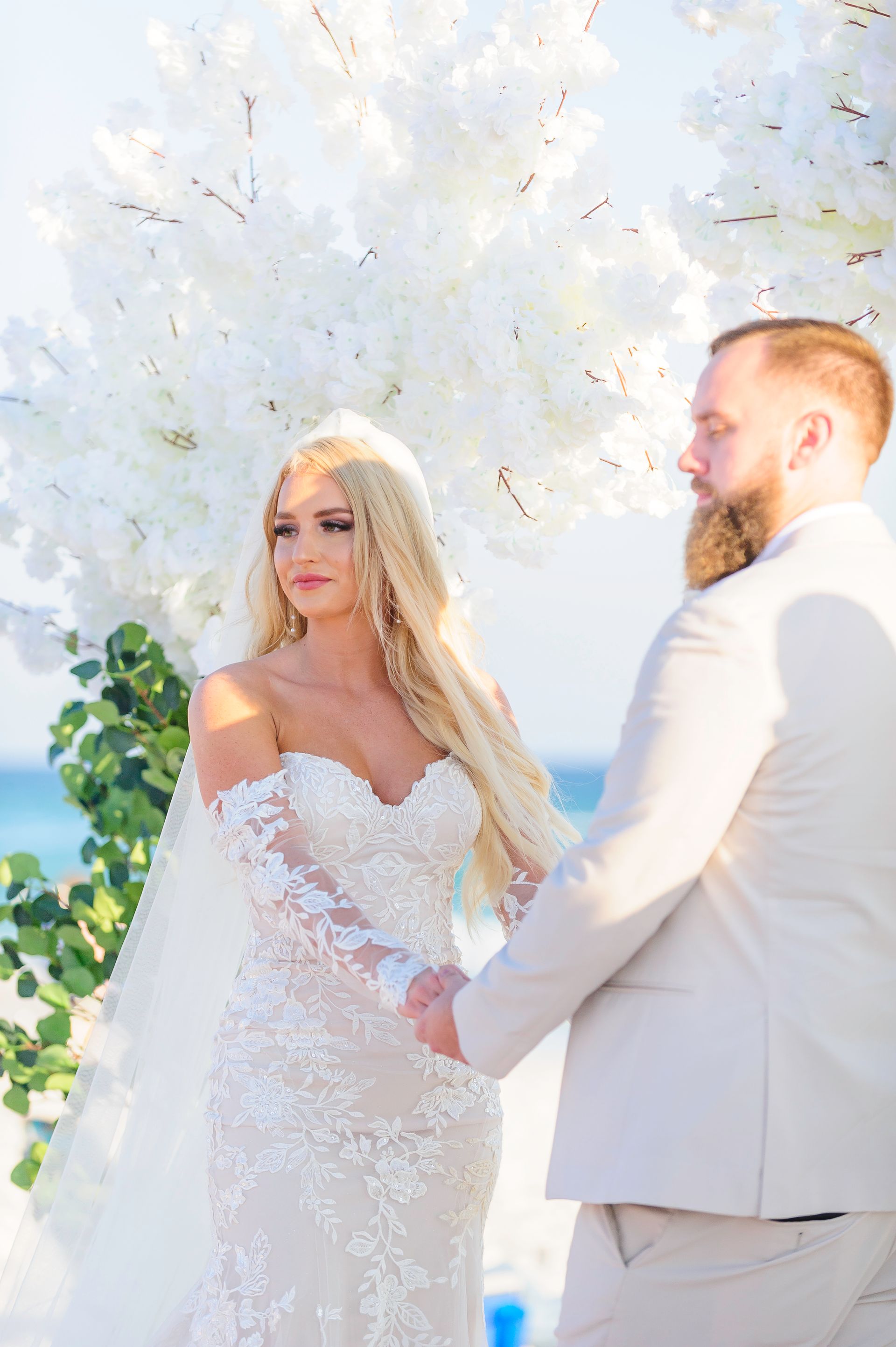 Bride and groom in white attire holding hands at a bright outdoor wedding ceremony