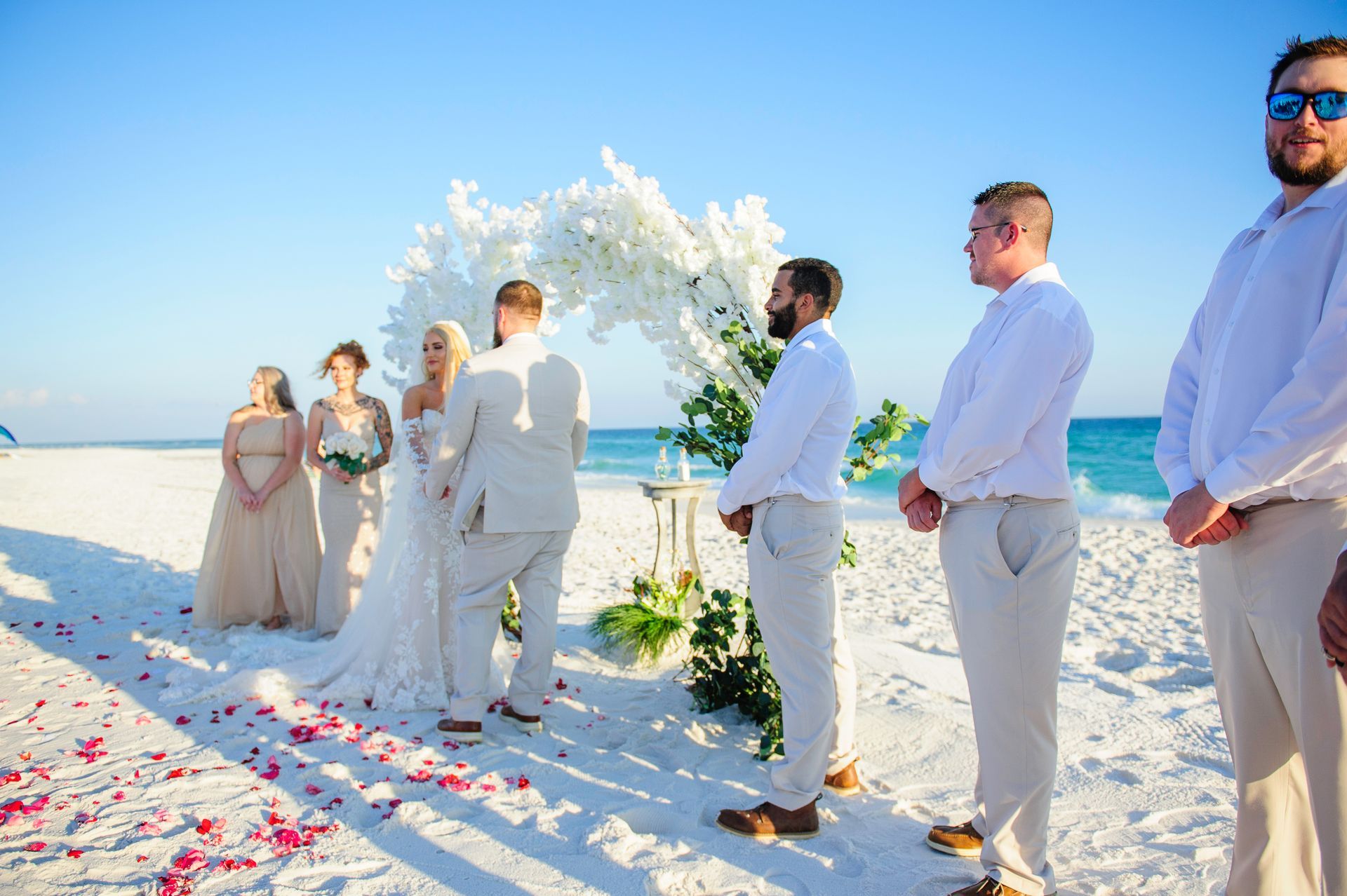 Wedding party standing on a flower-lined beach aisle by the ocean under a white floral arch