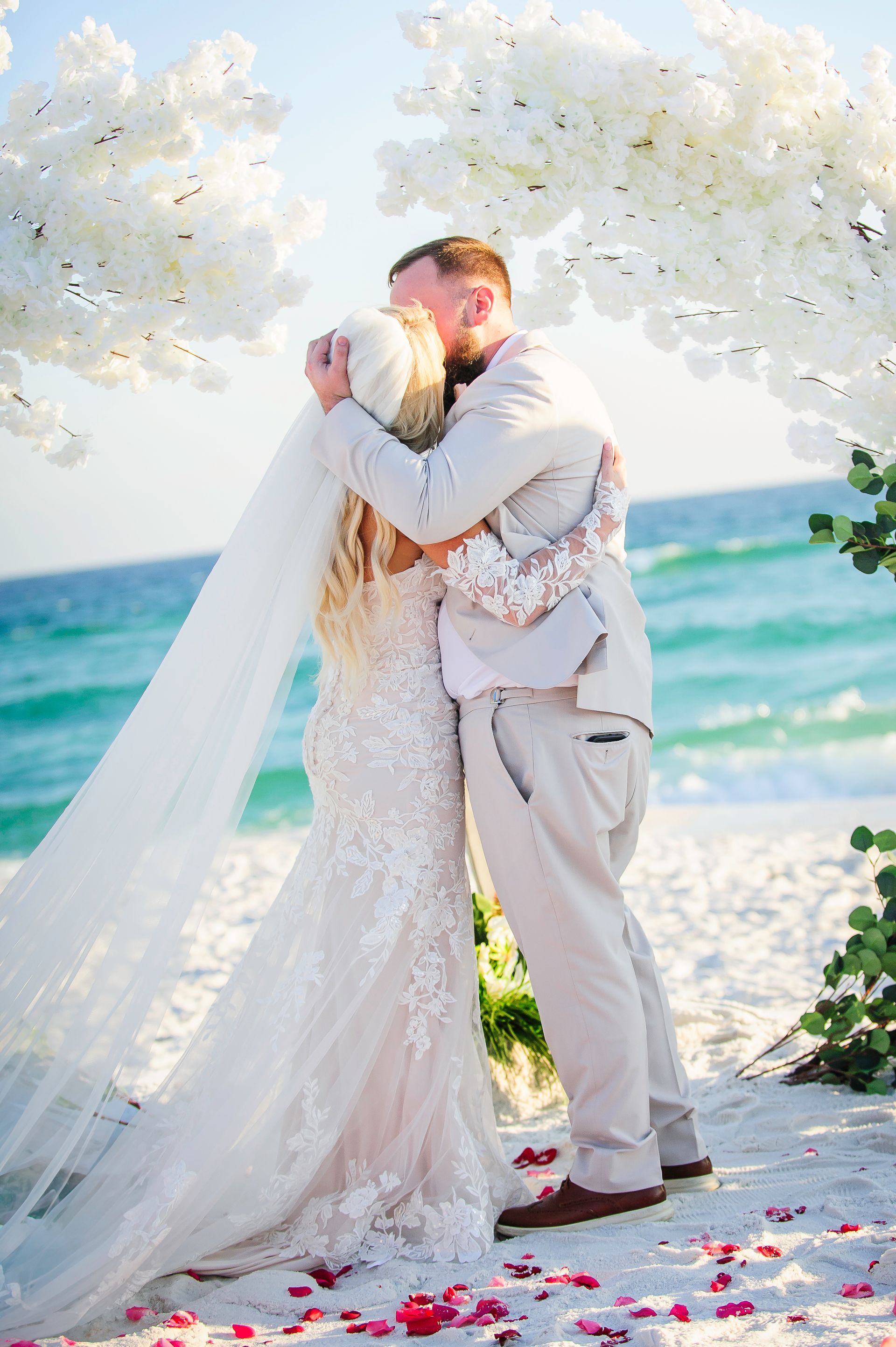 Couple embracing on a beach wedding arch with white flowers, ocean in the background and rose petals on the ground