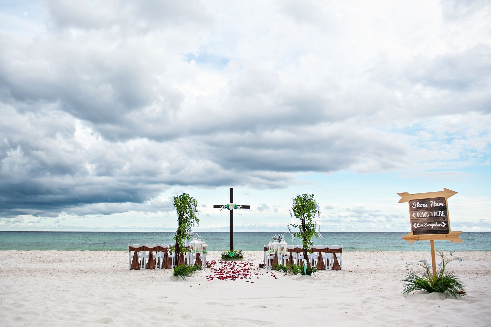 Beach wedding altar with white flowers, cross, chairs, and sign under cloudy sky by the sea