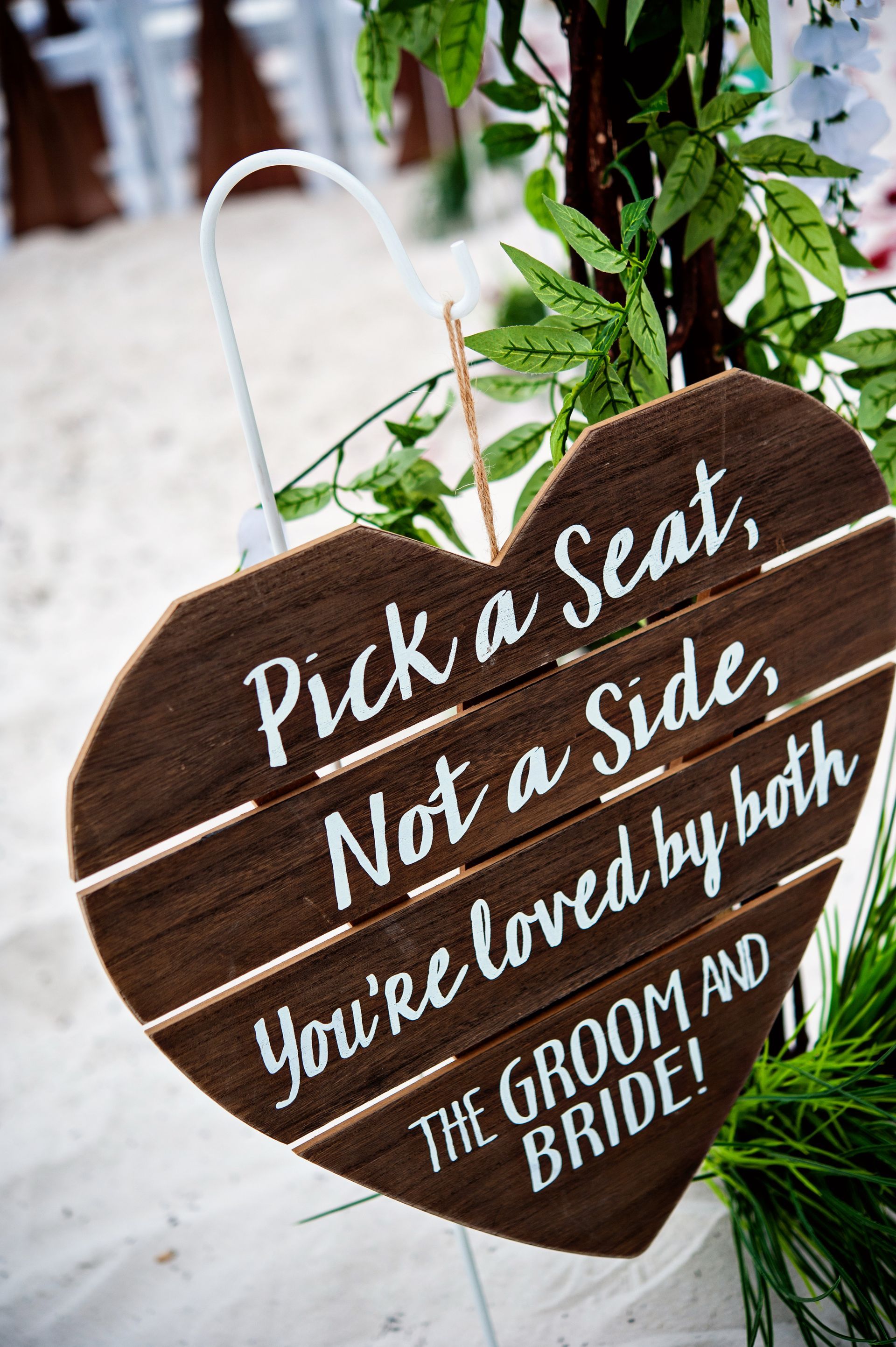 Heart-shaped wooden wedding sign reads, “Pick a seat, not a side. You’re loved by both the groom and bride.”