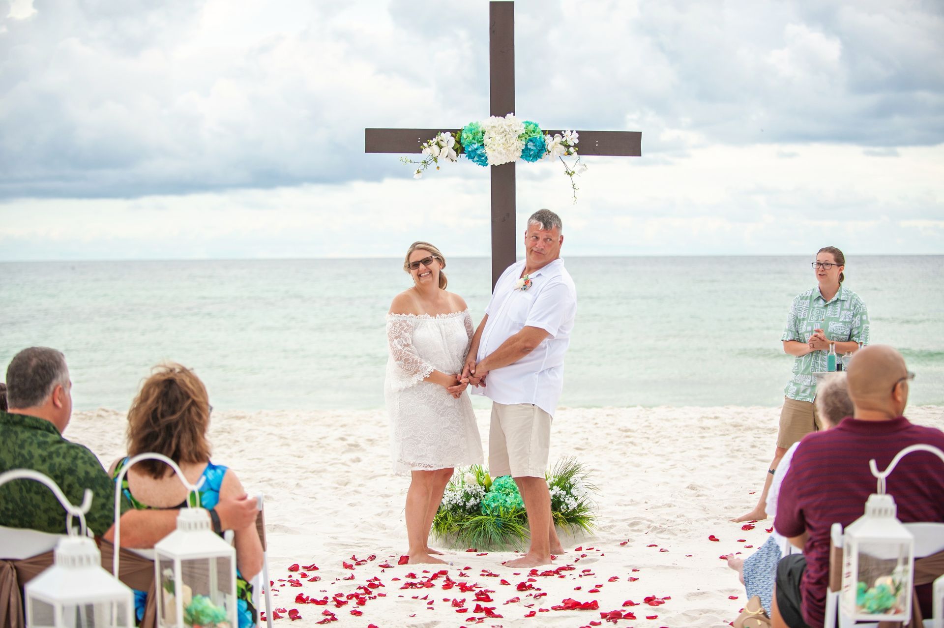 Beach wedding ceremony under a wooden cross, with guests seated on white chairs and flower petals on the sand.