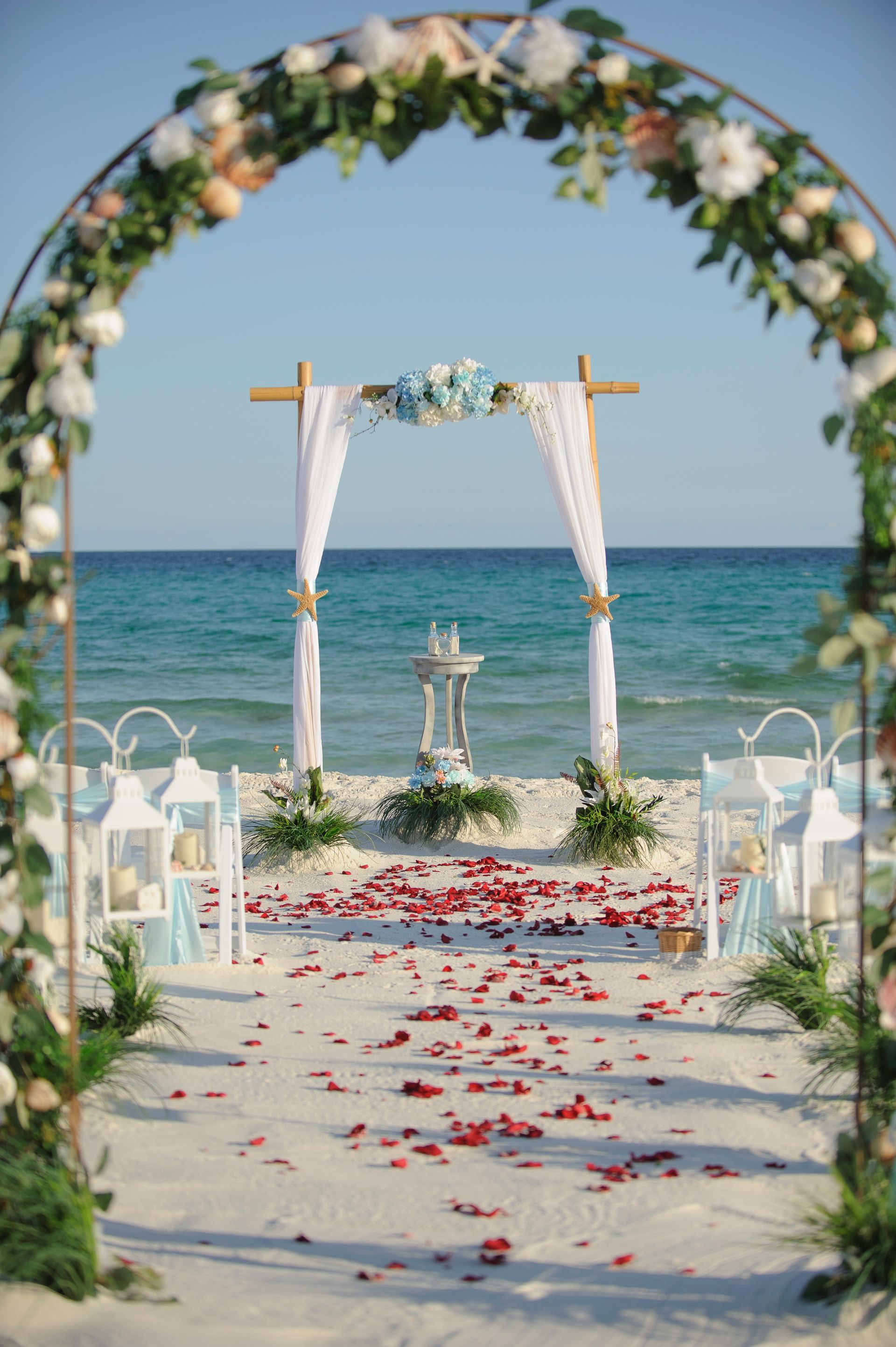 Beach wedding arch with white drapery and flowers facing the ocean, red petals scattered on the aisle