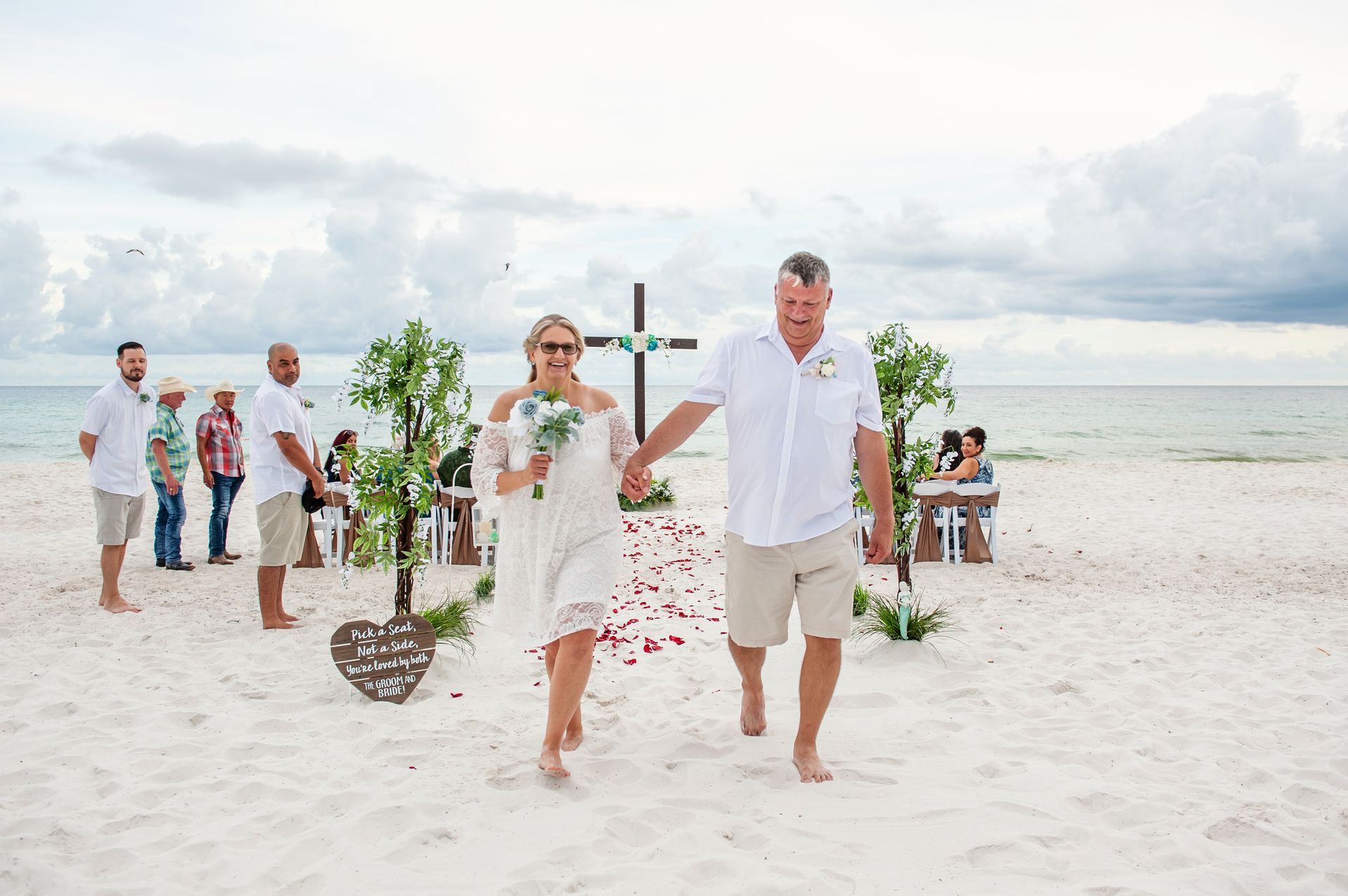 Couple walking barefoot down a white sandy beach wedding aisle, guests standing nearby by the ocean
