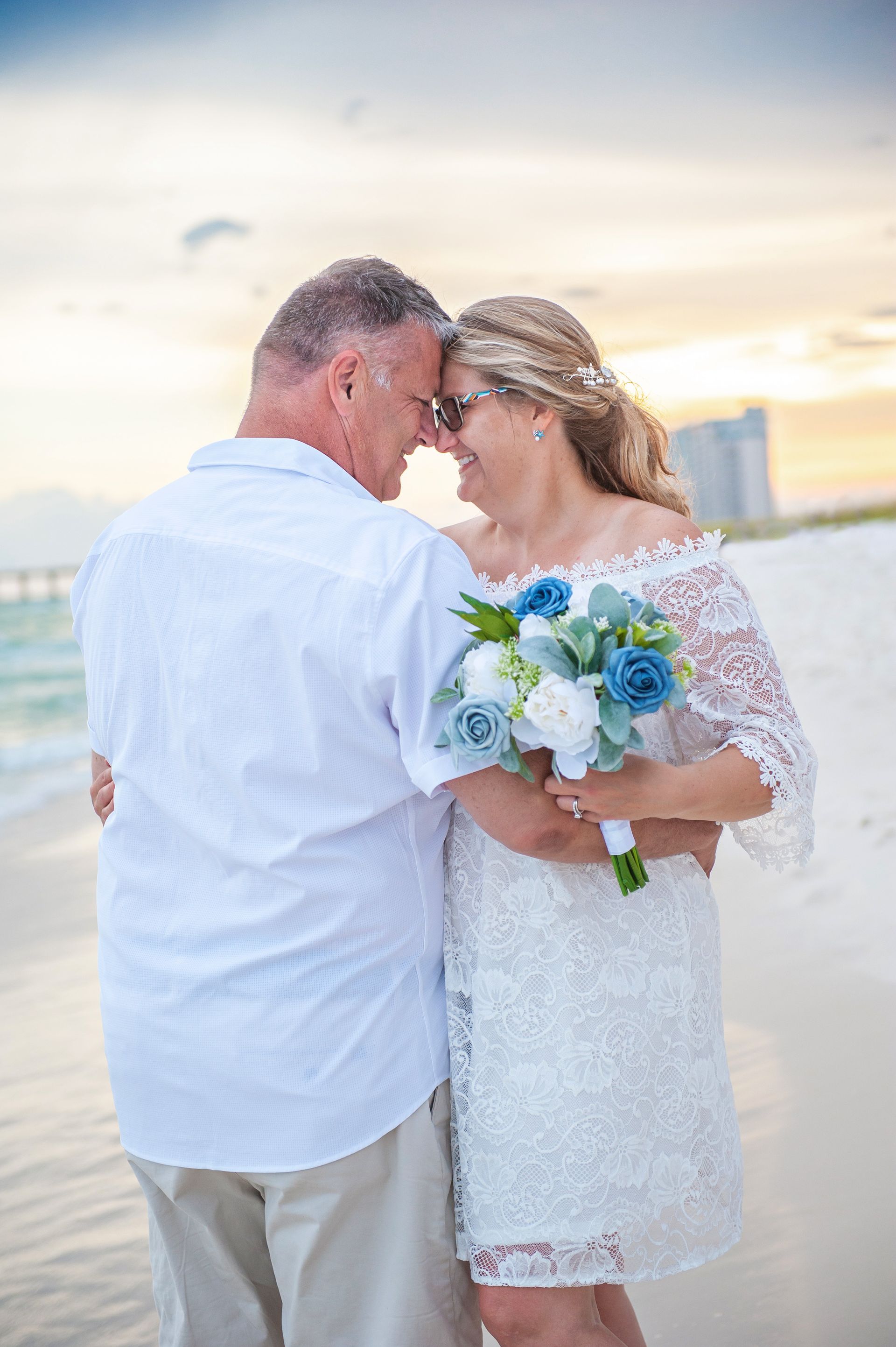Couple in white wedding attire sharing a tender moment by the beach at sunset, holding a bouquet