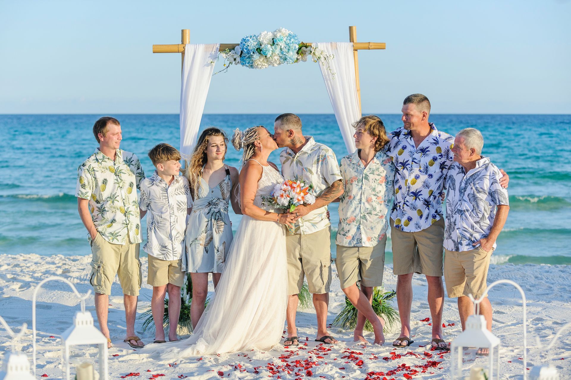 Beach wedding group under white arch, bride and groom holding bouquets by the ocean