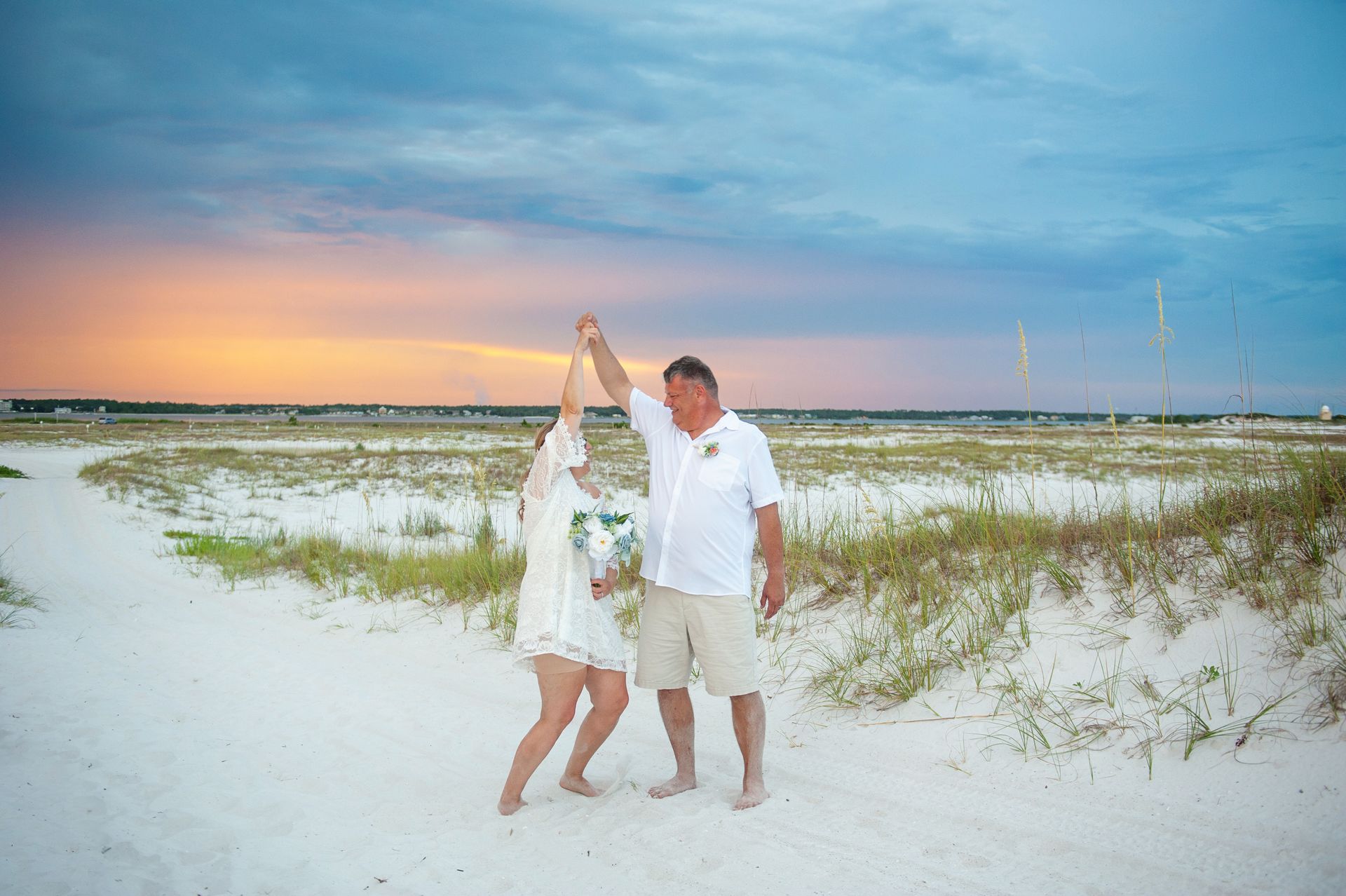 Couple holding hands on a beach at sunset, with grassy dunes and a pastel sky