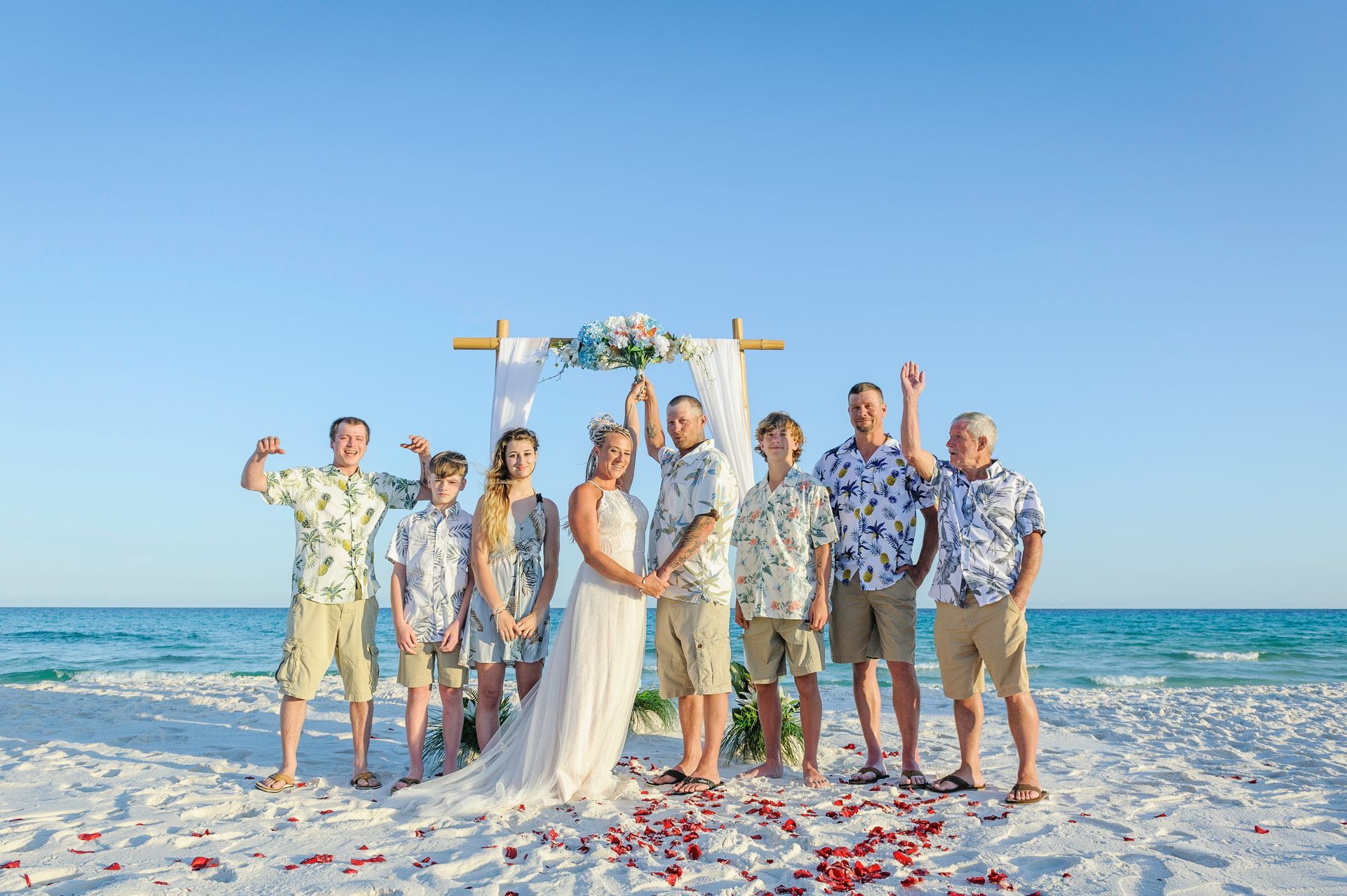 Wedding party posing on a sunny beach with ocean behind them, smiling amid confetti