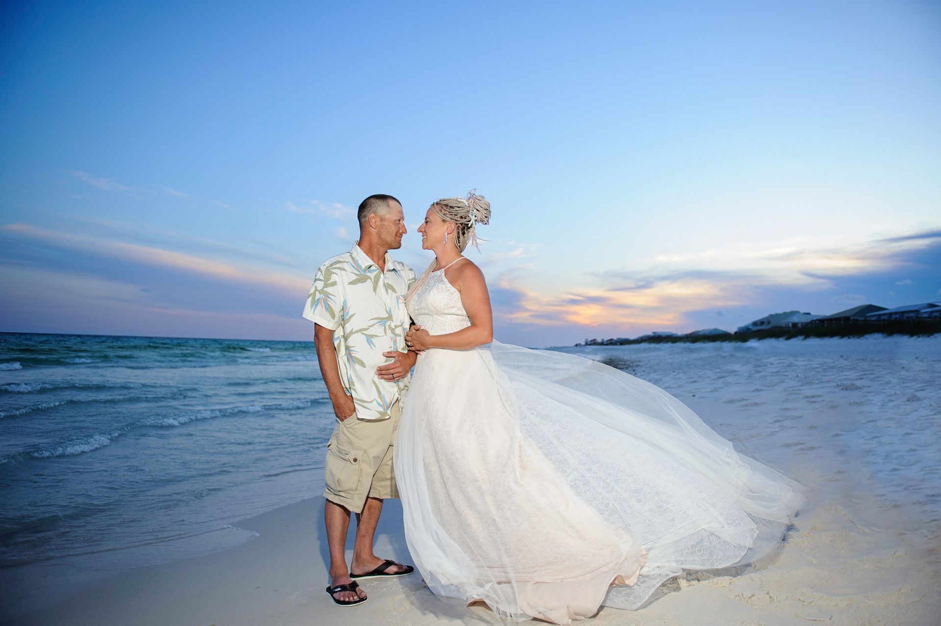 Couple in wedding attire standing on a beach at sunset, the bride’s dress flowing in the breeze.