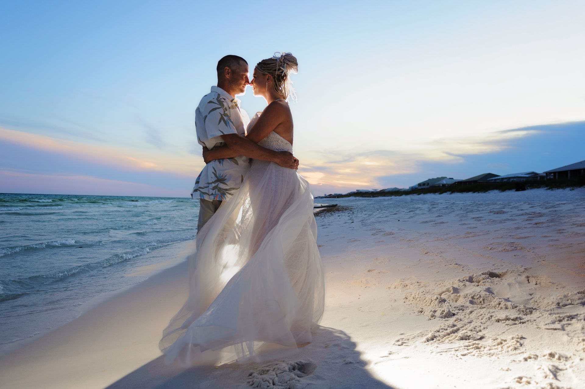 Couple embracing on a beach at sunset, wearing flowing white outfits with ocean waves in the background