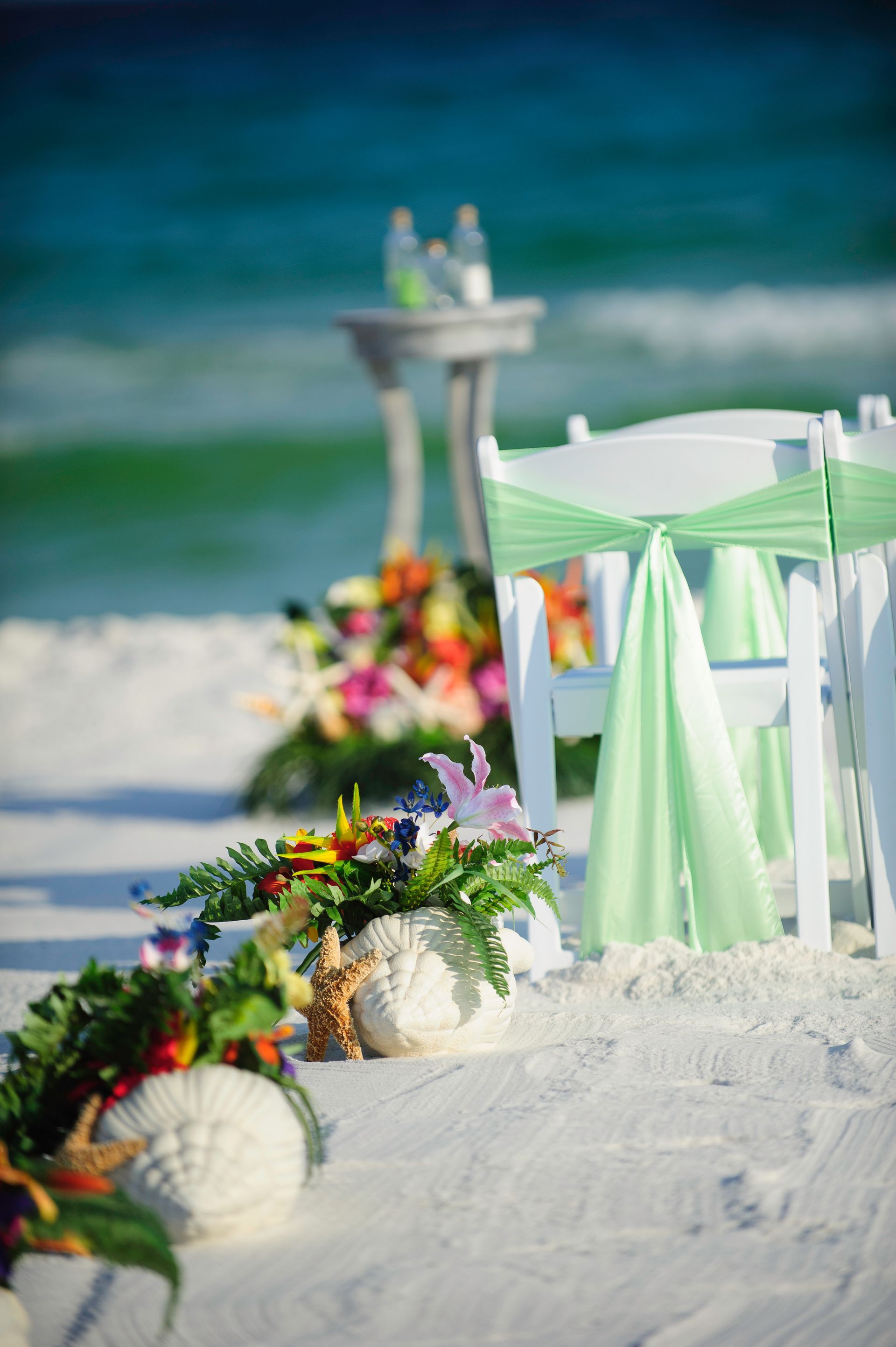Beach wedding setup with mint-green chairs, white aisle, and floral arrangements on the sand.