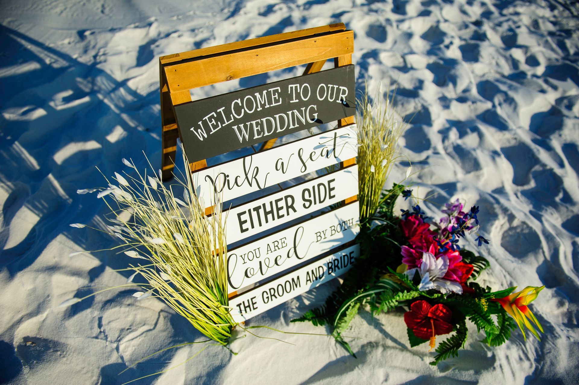 Wedding sign in the sand with flowers and seashells, reading “Welcome to our wedding” and “either side”