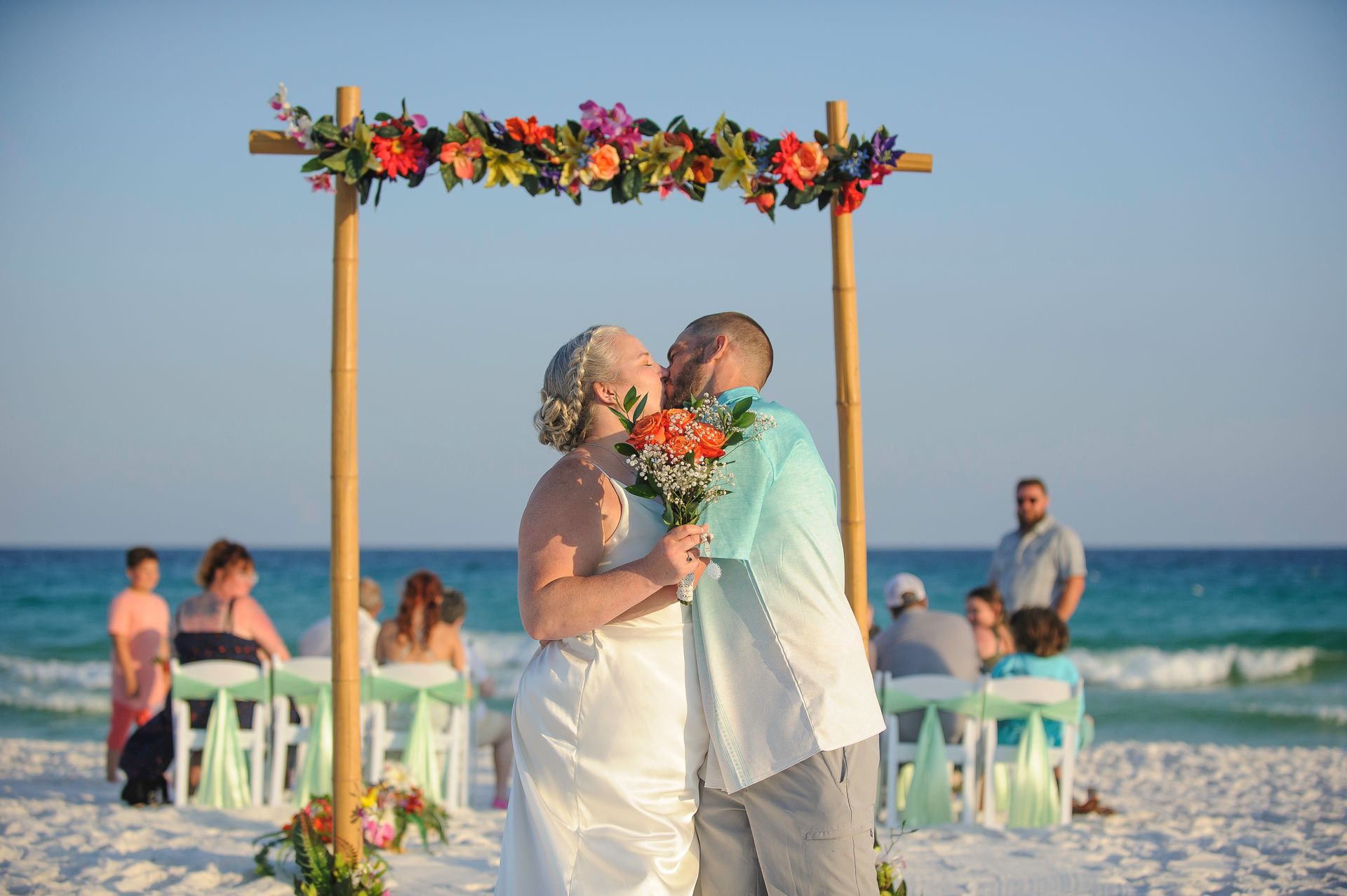 Couple exchanging vows under a flower arch on a beach, with seated guests and ocean waves in the background