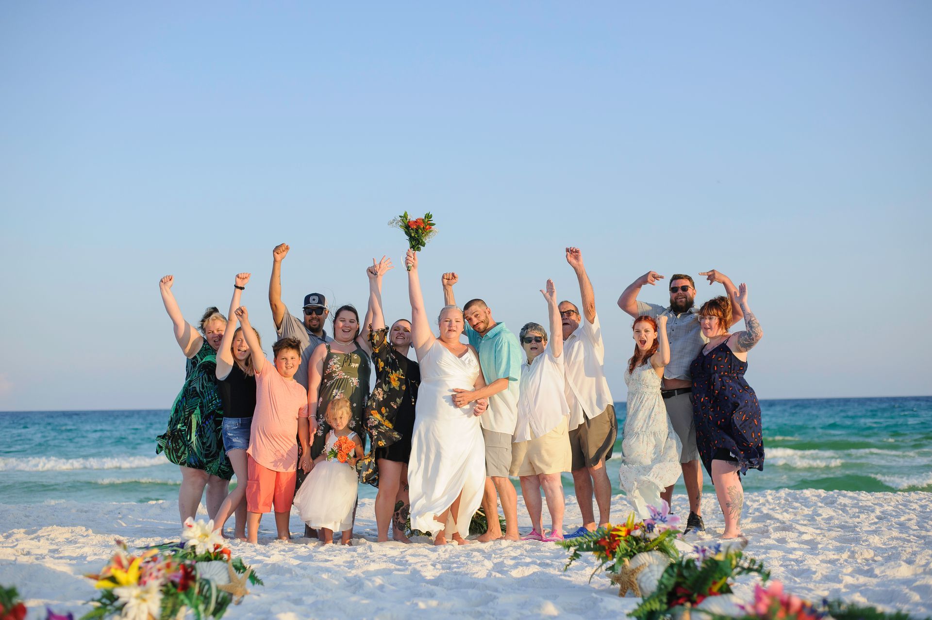 Group of people in wedding attire celebrating on a beach, arms raised beside the ocean.