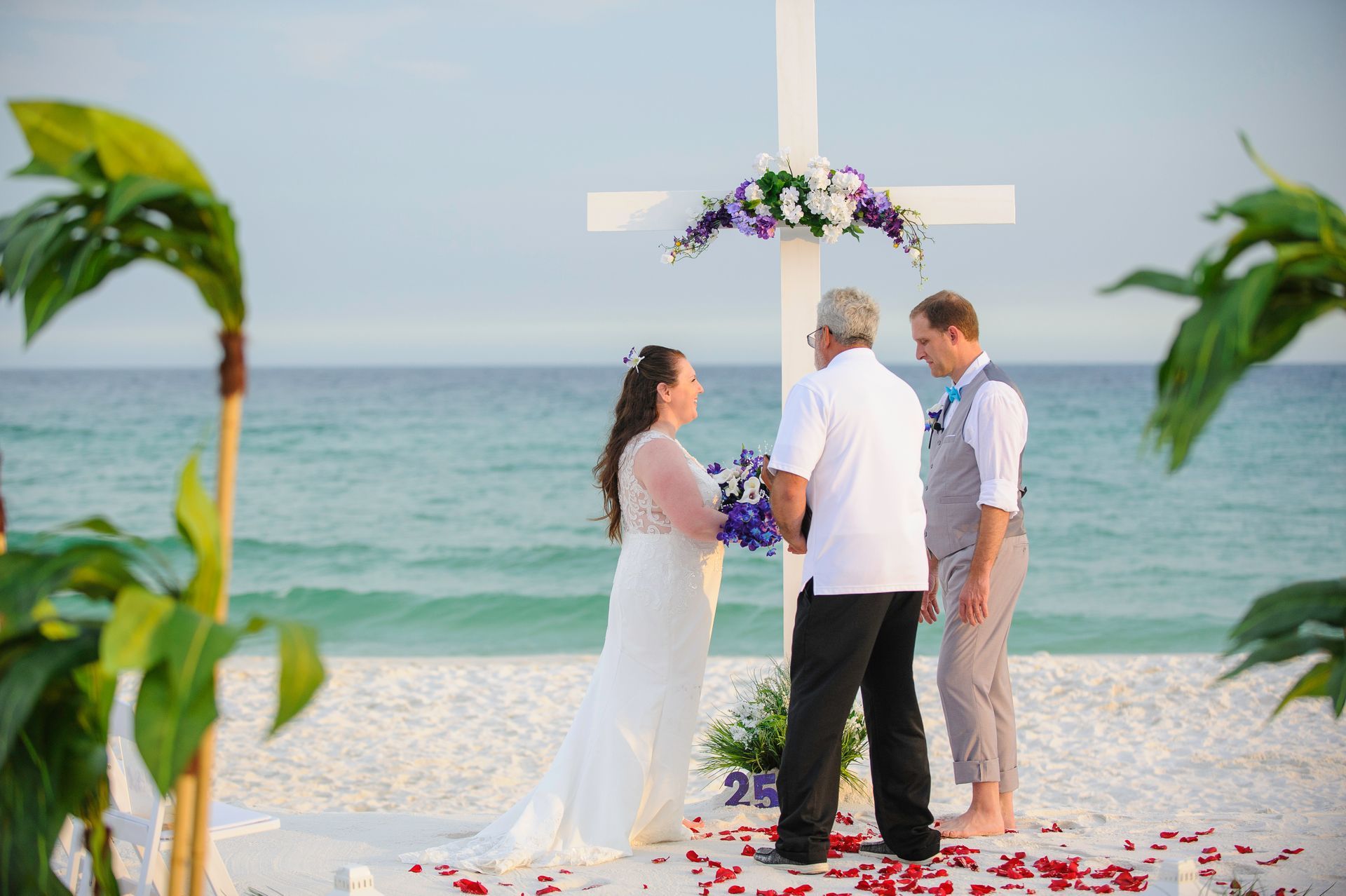 Beach wedding ceremony beneath a white cross arch with flowers, ocean in the background