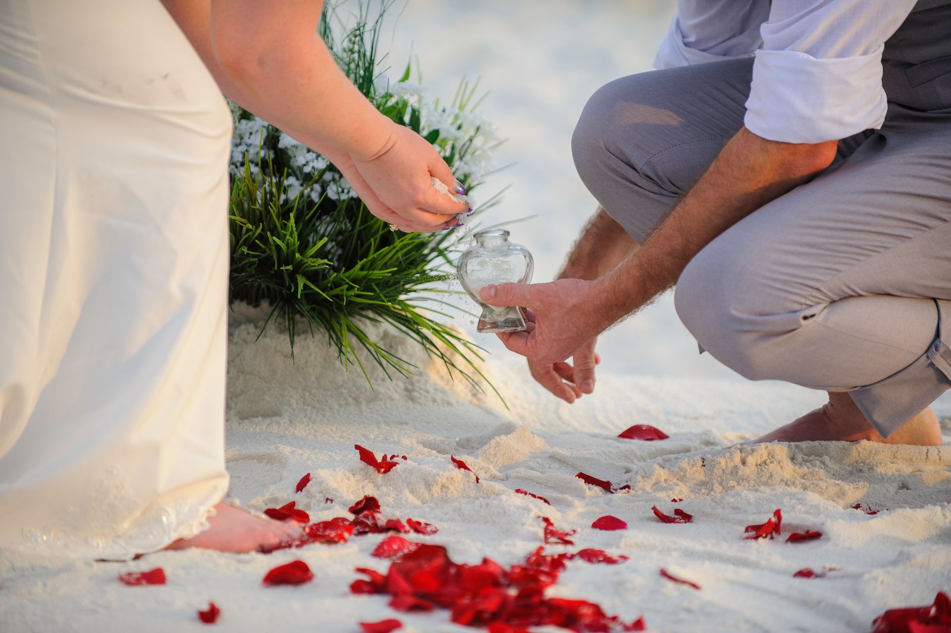 Two people kneel on a white sandy beach, pouring water over red flower petals in a ceremonial setting.