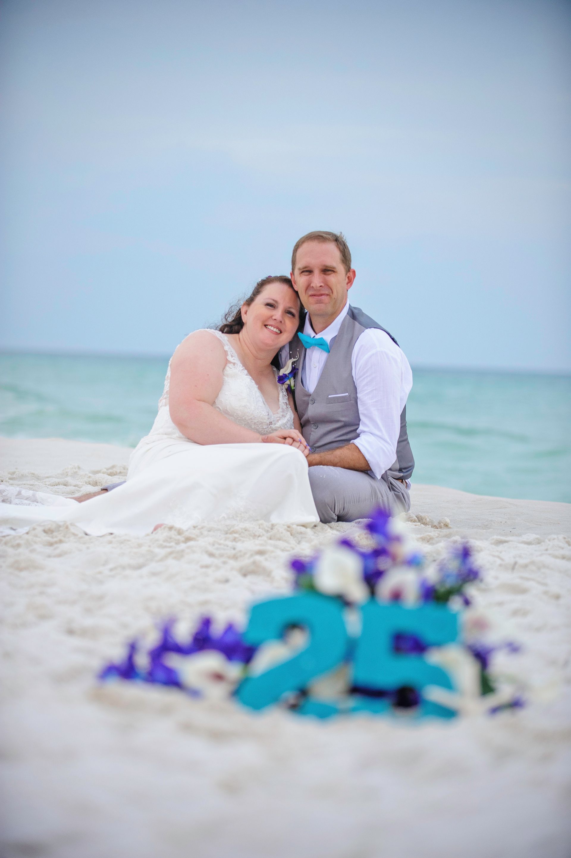 Couple in wedding attire sitting on a beach with turquoise ocean and blue flowers in the foreground