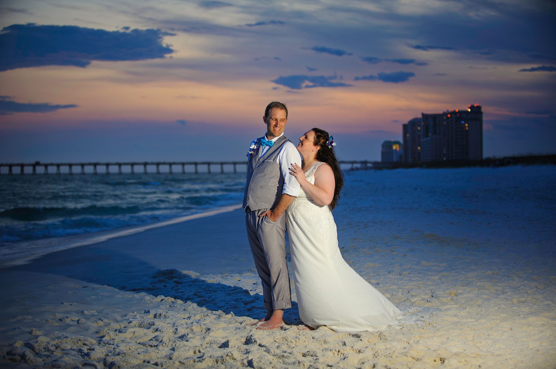 Newlyweds on a beach at sunset, with ocean waves and a pier in the background